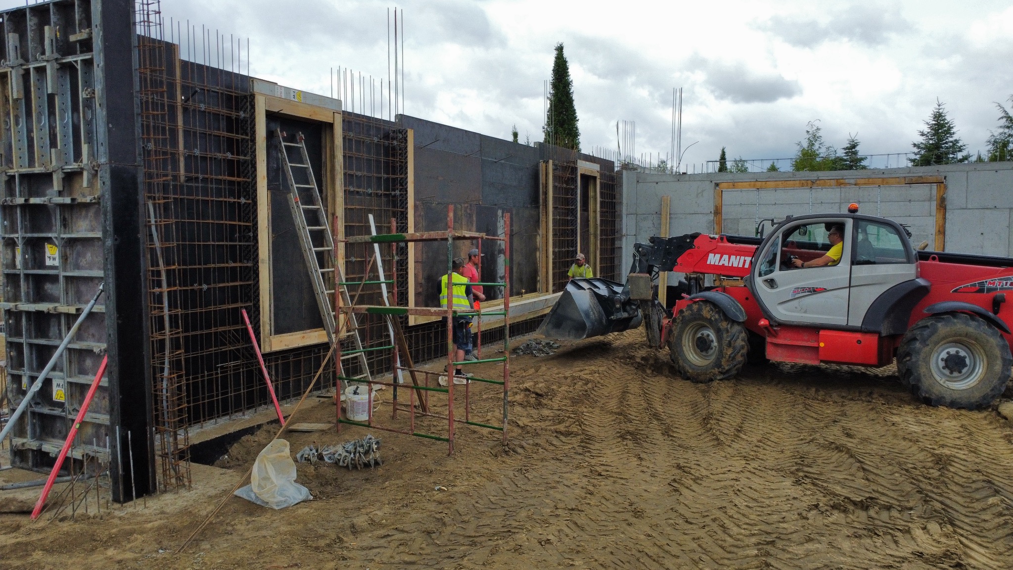 Budowa fundamentów z użyciem szalunków i zbrojenia, widoczny operator koparki Manitou. Prace ziemne w toku, teren budowy z rusztowaniami i materiałami.