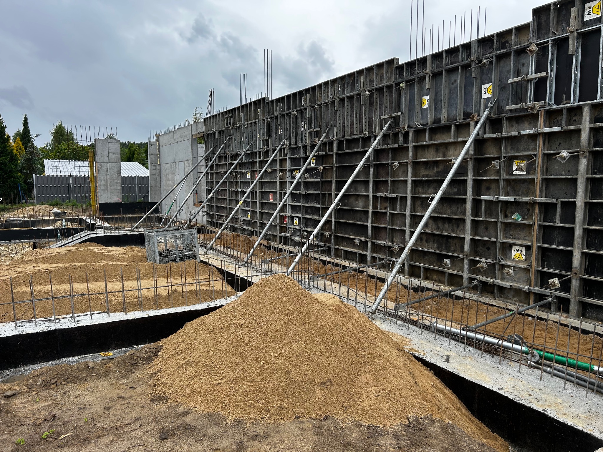 Szare szalunki z metalowymi podporami, świeży beton i piasek. Widok na budowę muru oporowego w pochmurny dzień. Konstrukcja zbrojona, przygotowana do zalania betonem.