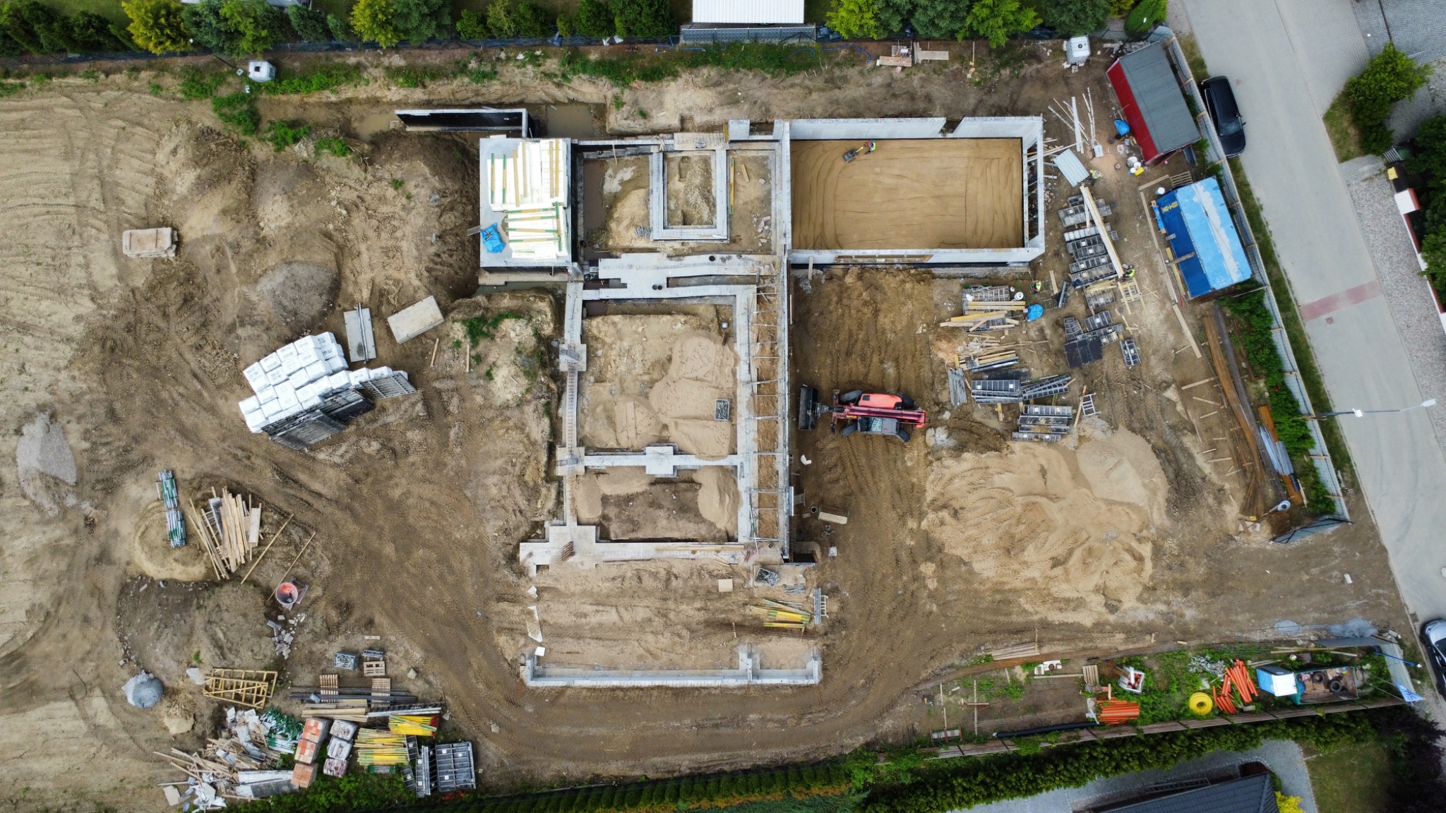 Widok z lotu ptaka na fundamenty domu w trakcie budowy. Betonowe ściany, piasek, sprzęt budowlany i materiały składowane na placu budowy.