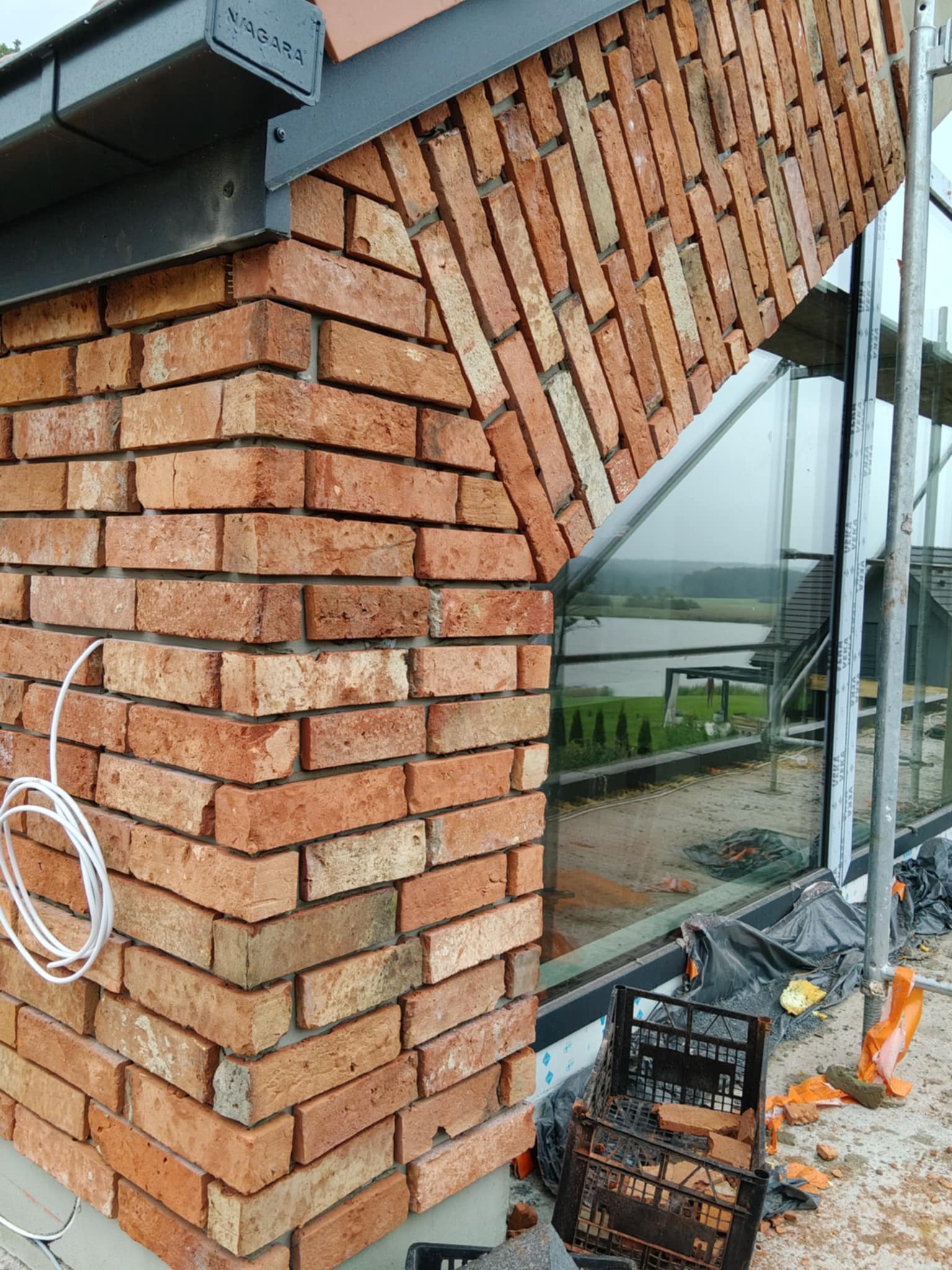 Fragment murowanej ściany z czerwonej cegły, z widocznym oknem i elementami rusztowania. Nietypowe ułożenie cegieł tworzy łuk nad oknem. Widoczne elementy wykończeniowe.