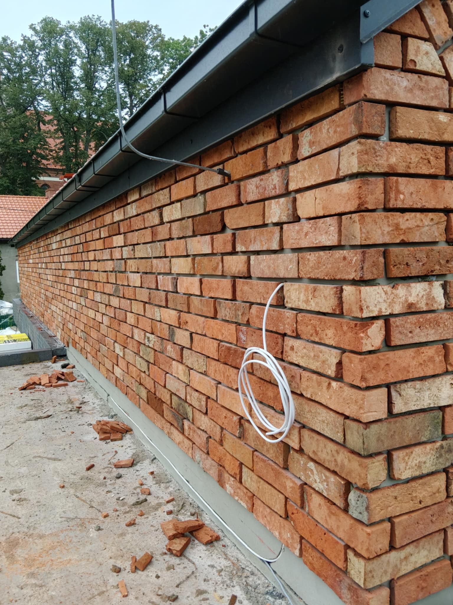 Ściana z czerwonej cegły klinkierowej pod metalowym dachem, z widocznym przewodem elektrycznym i drobnymi fragmentami cegieł na posadzce.
