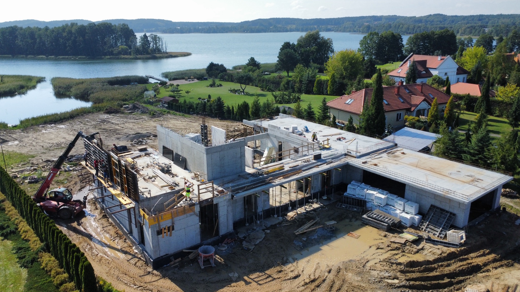 Budowa domu w stanie surowym otwartym, widok z góry na betonowe ściany i fundamenty. Na budowie obecny żuraw, w tle jezioro i zabudowa mieszkalna.