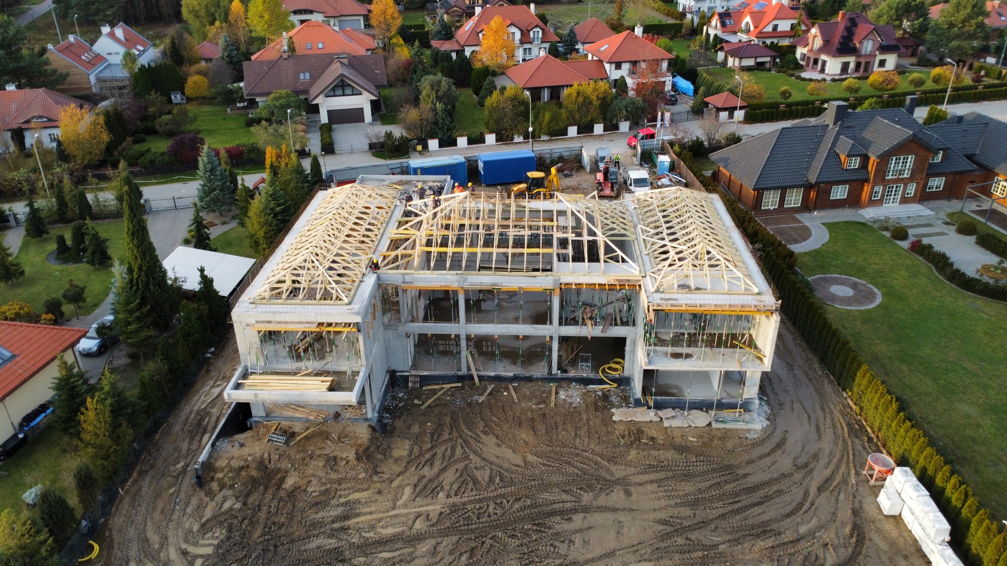 Budowa domu jednorodzinnego w stanie surowym otwartym z drewnianą konstrukcją dachu, widok z lotu ptaka na tle zabudowy mieszkalnej i zieleni.