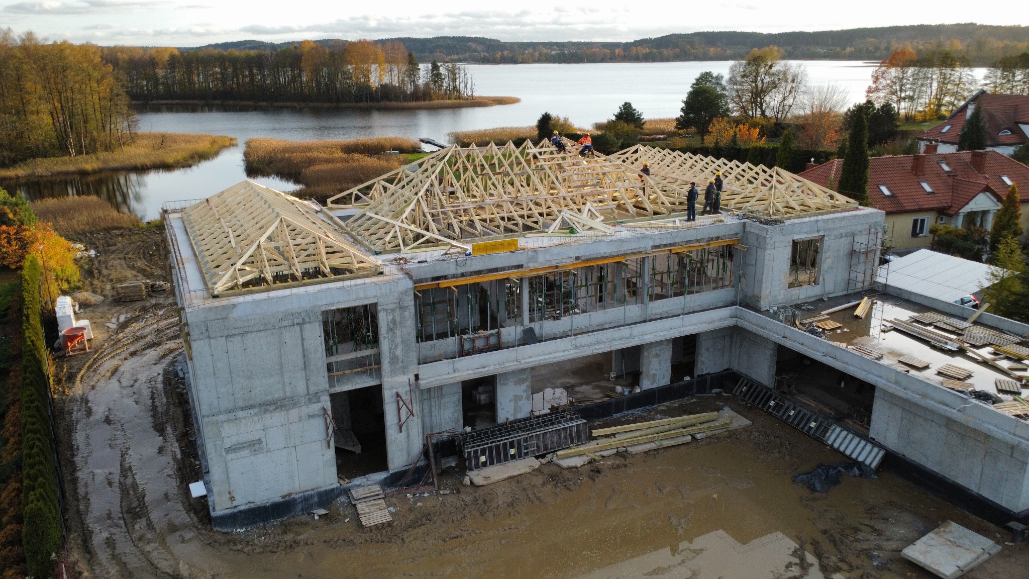 Budowa domu w stanie surowym otwartym z drewnianą konstrukcją dachu w trakcie montażu. W tle jezioro i jesienny krajobraz. Widok z lotu ptaka.