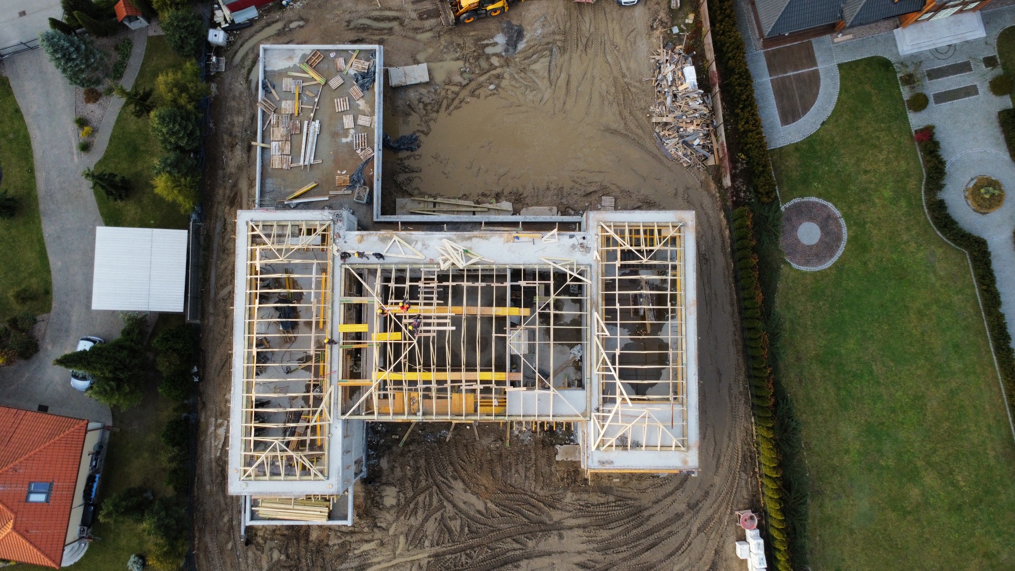 Zdjęcie z drona: budowa domu w trakcie wznoszenia więźby dachowej. Widoczne fundamenty, ściany i drewniana konstrukcja dachu. Na placu budowy pracownicy w kaskach.