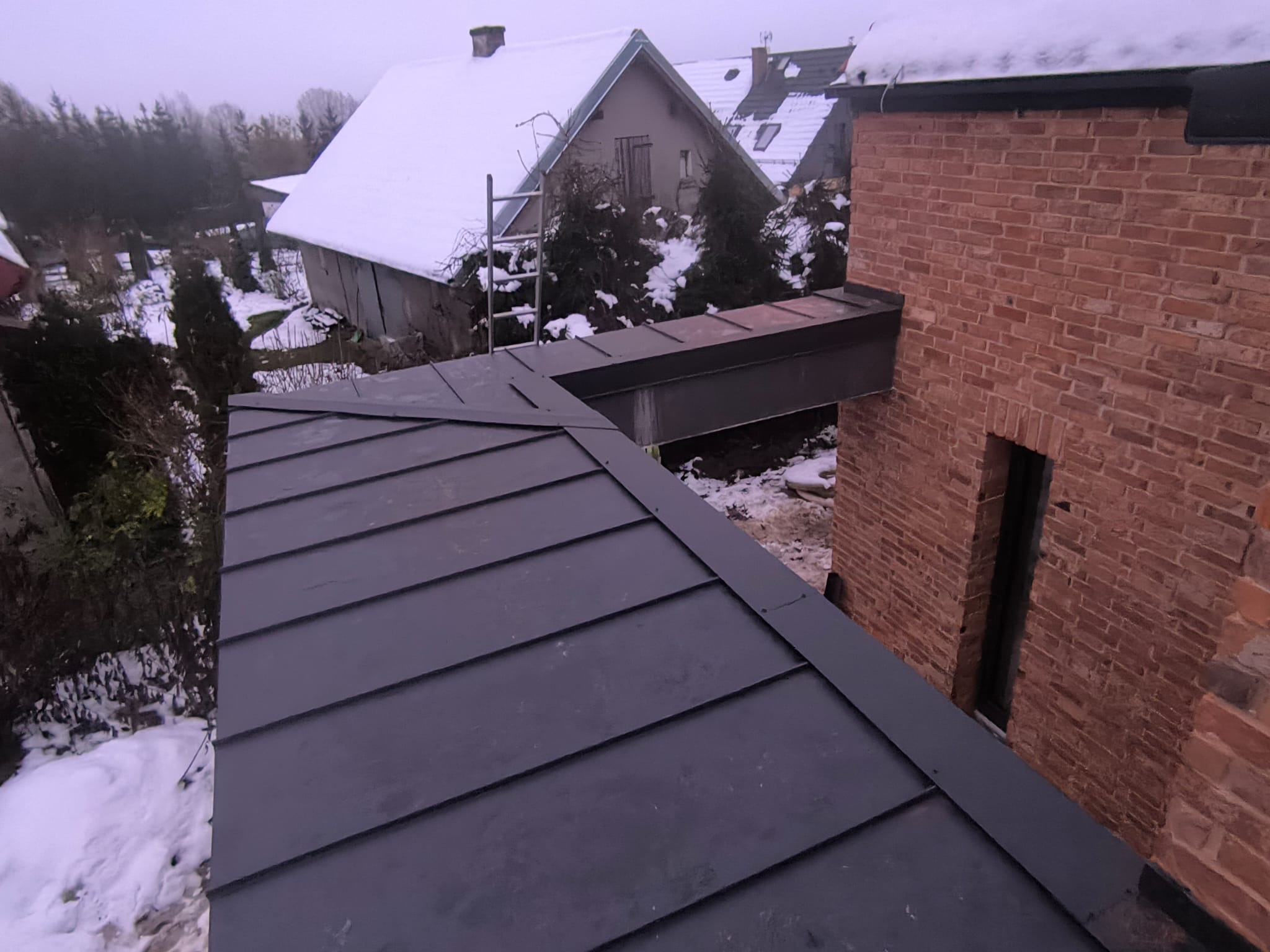 Nowy, grafitowy dach z blachy na domu z cegły, połączony stalową konstrukcją. W tle ośnieżone budynki i drzewa. Widok z góry, pochmurny dzień.