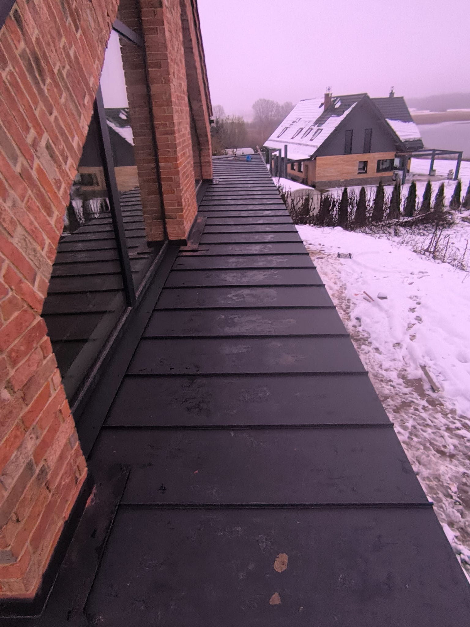 Nowoczesny dom z dachem z blachy na rąbek stojący, widok z góry na dach i komin z cegły klinkierowej, w tle zasypany śniegiem ogród i drugi budynek.