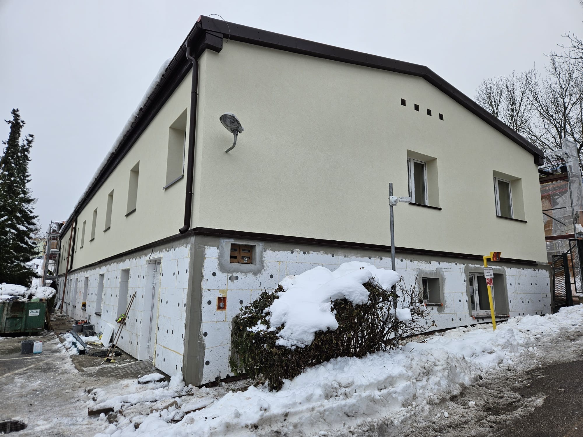Budynek w trakcie ocieplania styropianem, widoczne narzędzia i śnieg. Część budynku otynkowana na kremowo, z brązowym dachem. Prace w toku, zimowa sceneria.