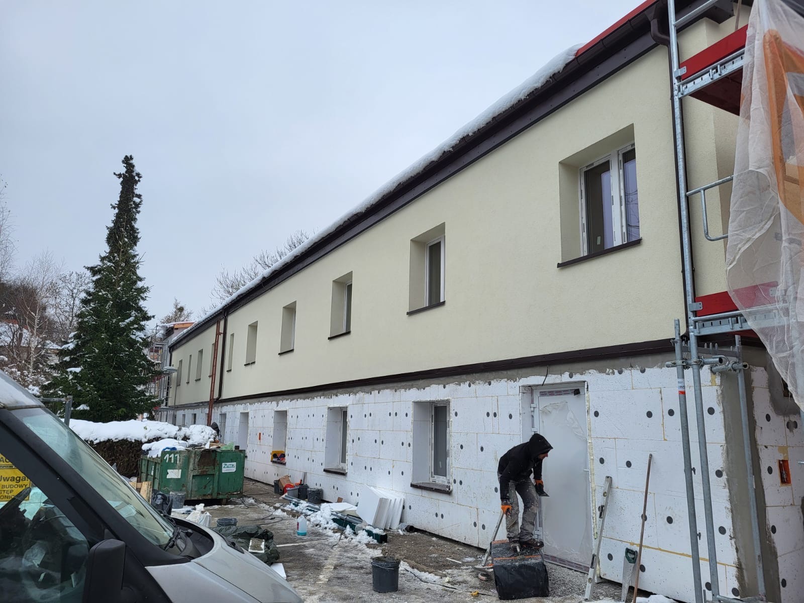 Ocieplanie budynku styropianem: pracownik na rusztowaniu przykłada arkusz do ściany. Widoczne okna, ośnieżone otoczenie i fragment samochodu.