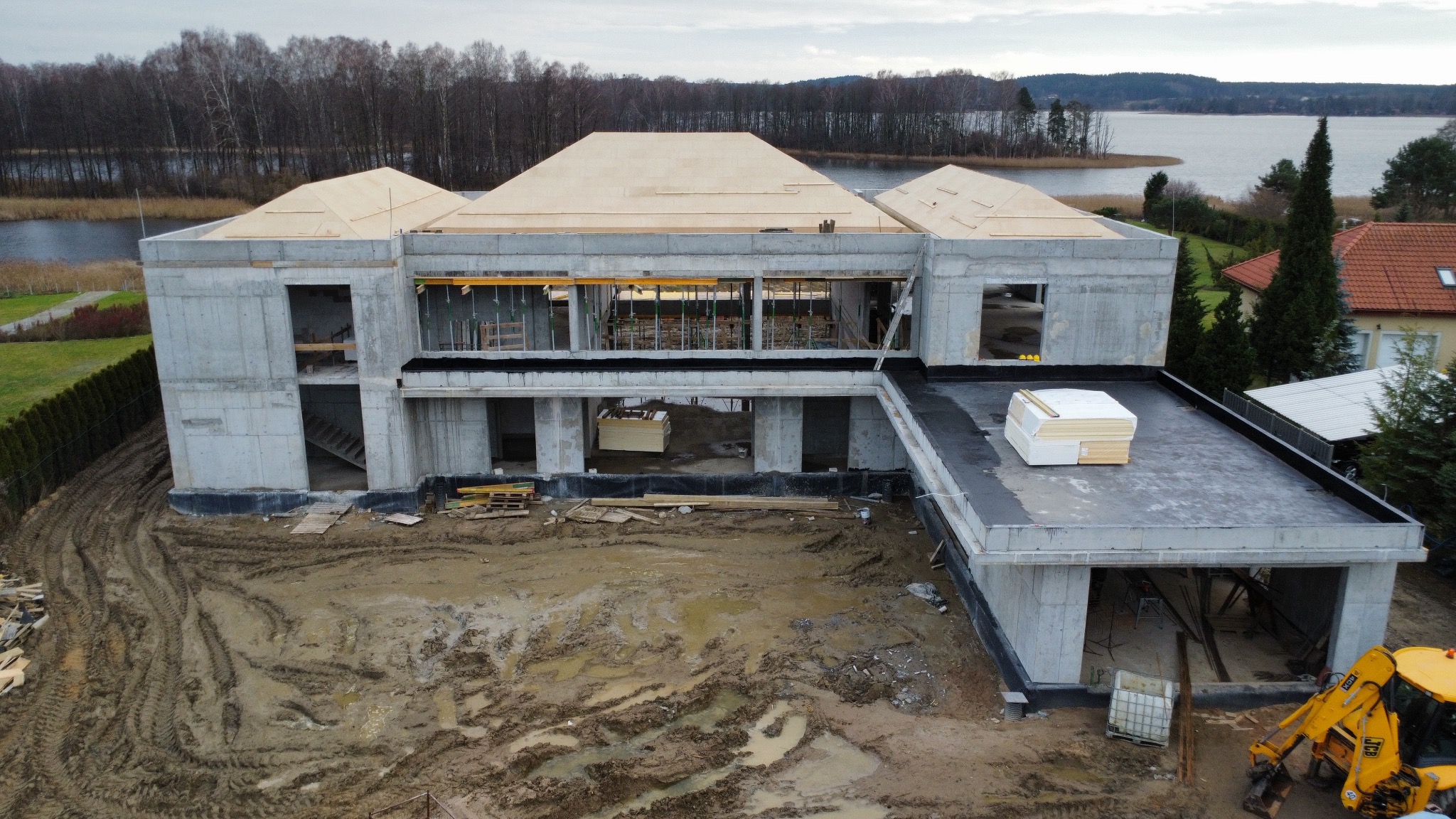 Surowy, dwukondygnacyjny dom z betonu w trakcie budowy, z widoczną drewnianą konstrukcją dachu i żółtą koparką na błotnistym terenie. W tle jezioro i drzewa.
