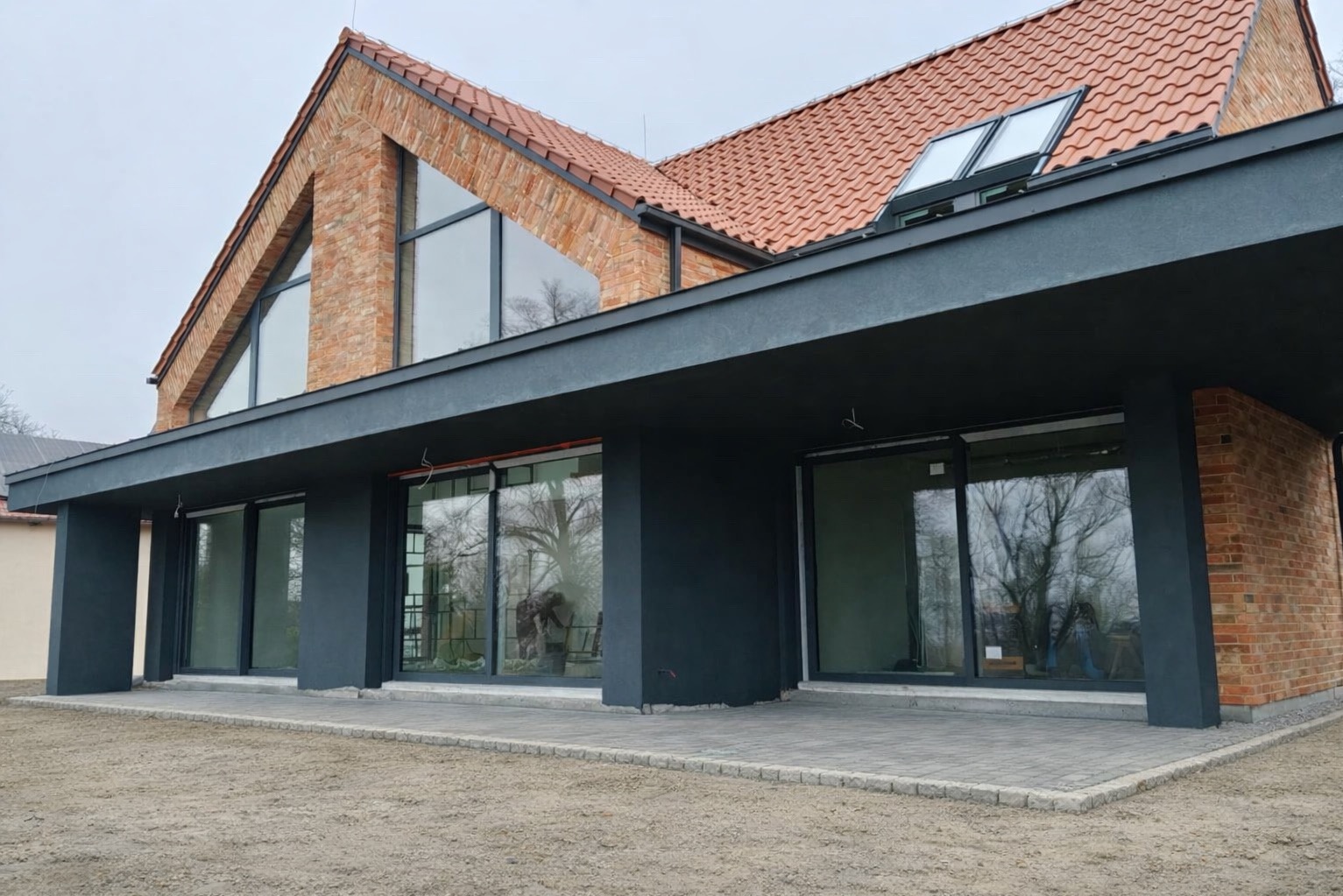 Nowoczesny dom z cegły klinkierowej w Olsztynie. Duże przeszklenia, dachówka ceramiczna, grafitowe elementy wykończeniowe. Widok z zewnątrz, w tle drzewa.