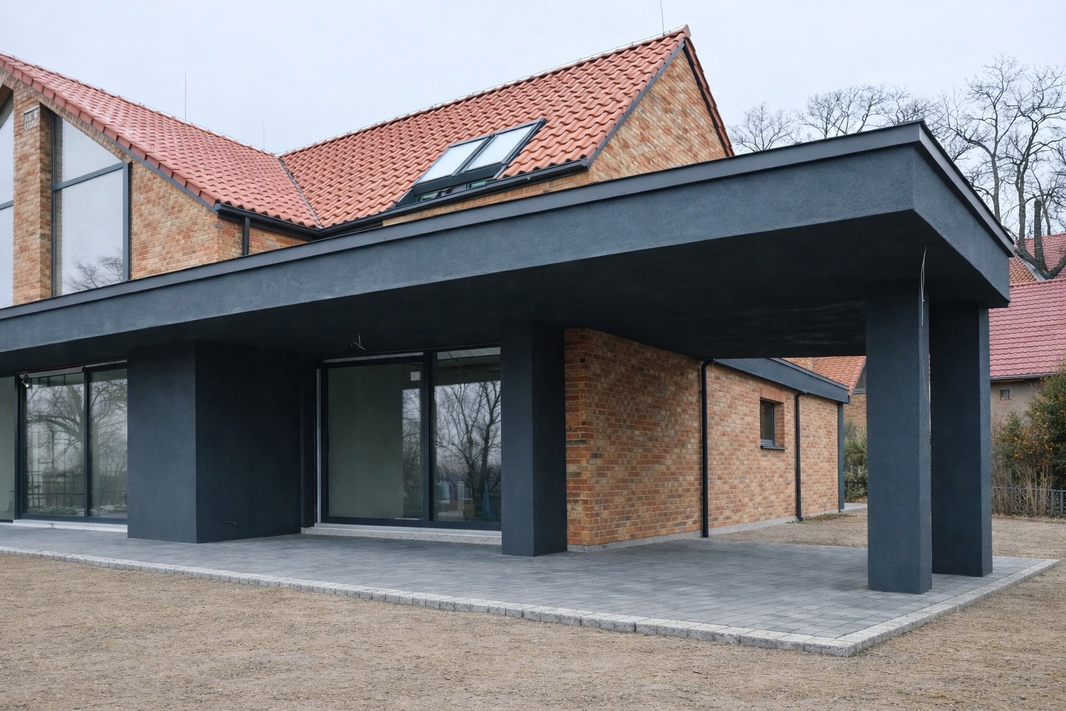 Nowoczesny dom z cegły klinkierowej z ciemnoszarą wiatą garażową i dużymi oknami przesuwnymi. Widoczny fragment dachu z dachówką i oknami połaciowymi.