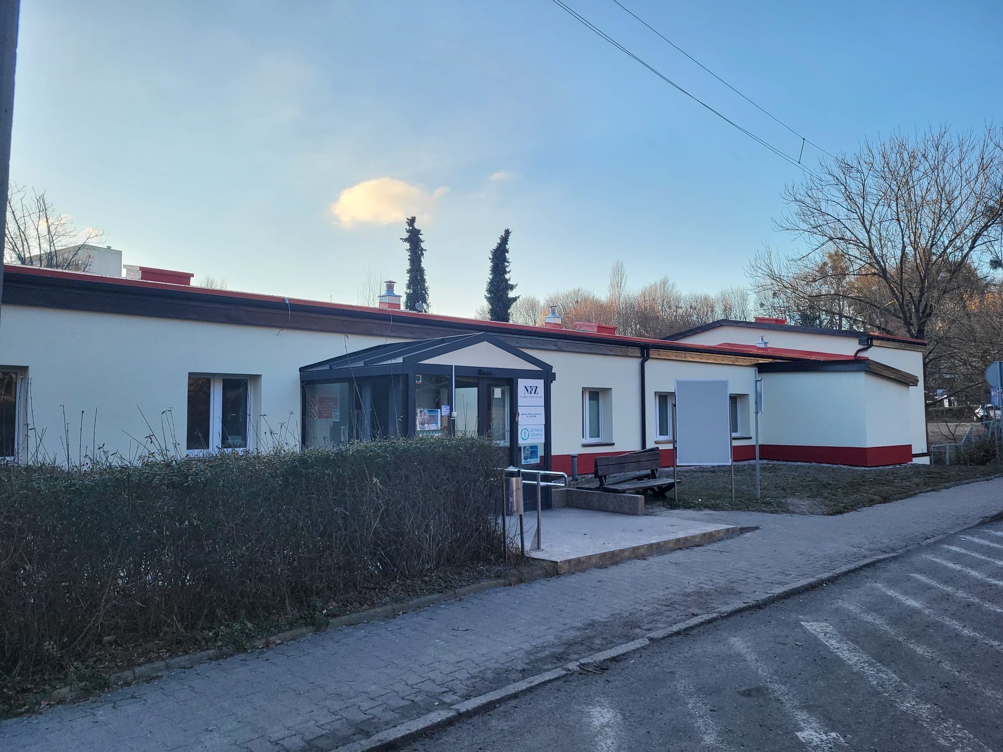 Parterowy budynek z przeszklonym wejściem i czerwonymi akcentami, otoczony zielenią. Widok z poziomu ulicy w słoneczny dzień.