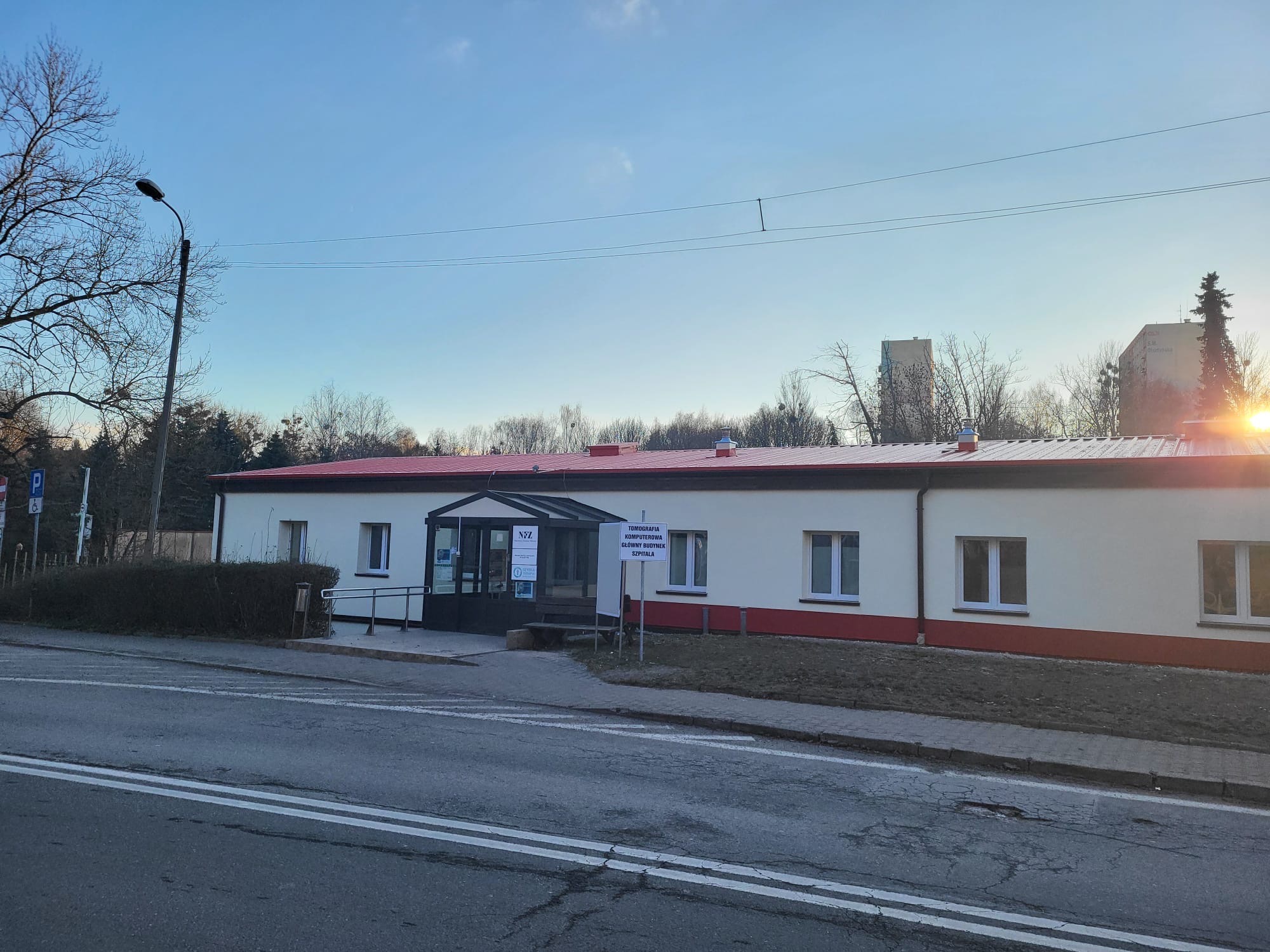 Parterowy budynek z czerwonym dachem i białymi ścianami, widoczny z ulicy w słoneczny dzień. Wejście z rampą dla niepełnosprawnych. Tabliczka informacyjna przed wejściem.