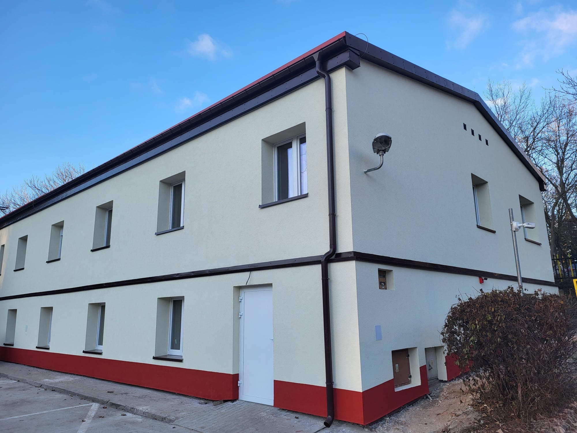 Dwukondygnacyjny budynek po remoncie elewacji, z nowymi oknami, pomalowany na biało z czerwonym pasem u dołu i brązowymi rynnami, oświetlony lampą zewnętrzną.