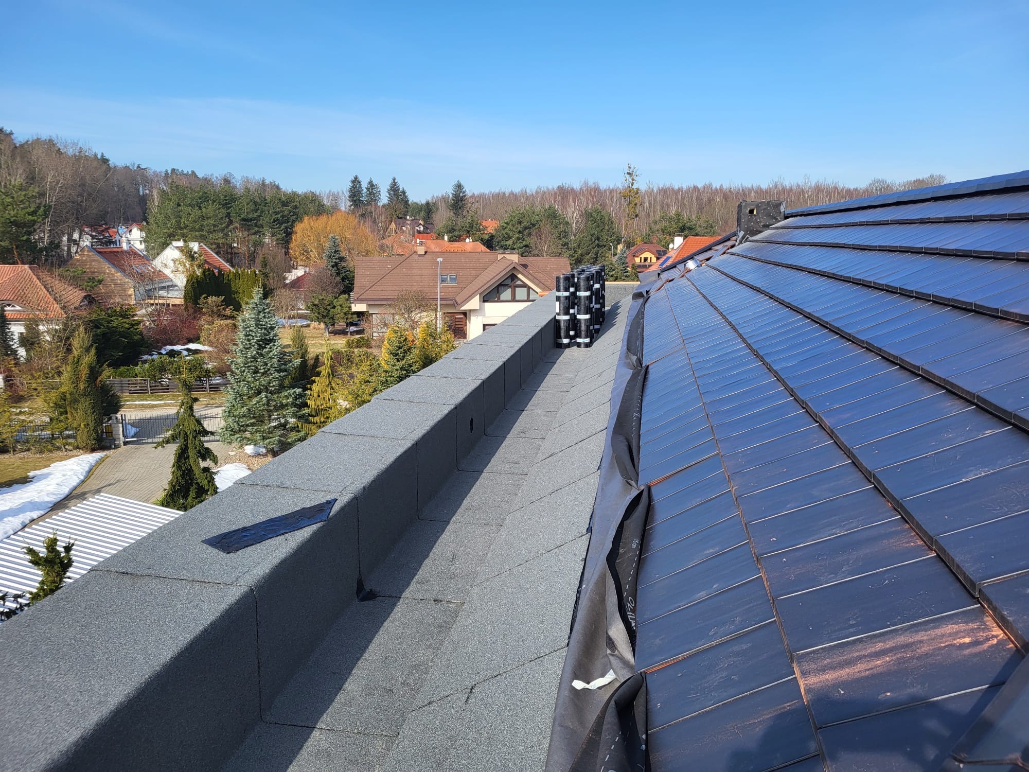 Nowy dach z ciemnej dachówki ceramicznej, widok z góry na tle zabudowy jednorodzinnej i lasu. Na dachu rolki papy. Słoneczny dzień.