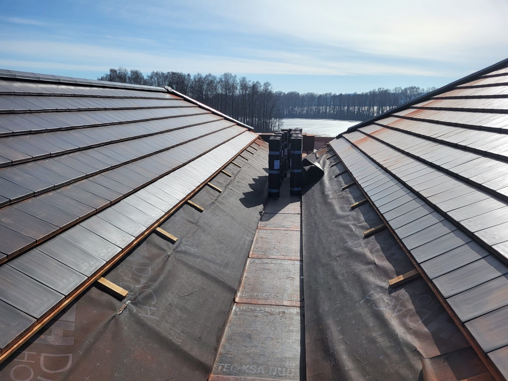 Układanie ciemnej dachówki ceramicznej na dachu domu, widok z góry na fragment dachu z kominem i membraną dachową, w tle błękitne niebo i drzewa.