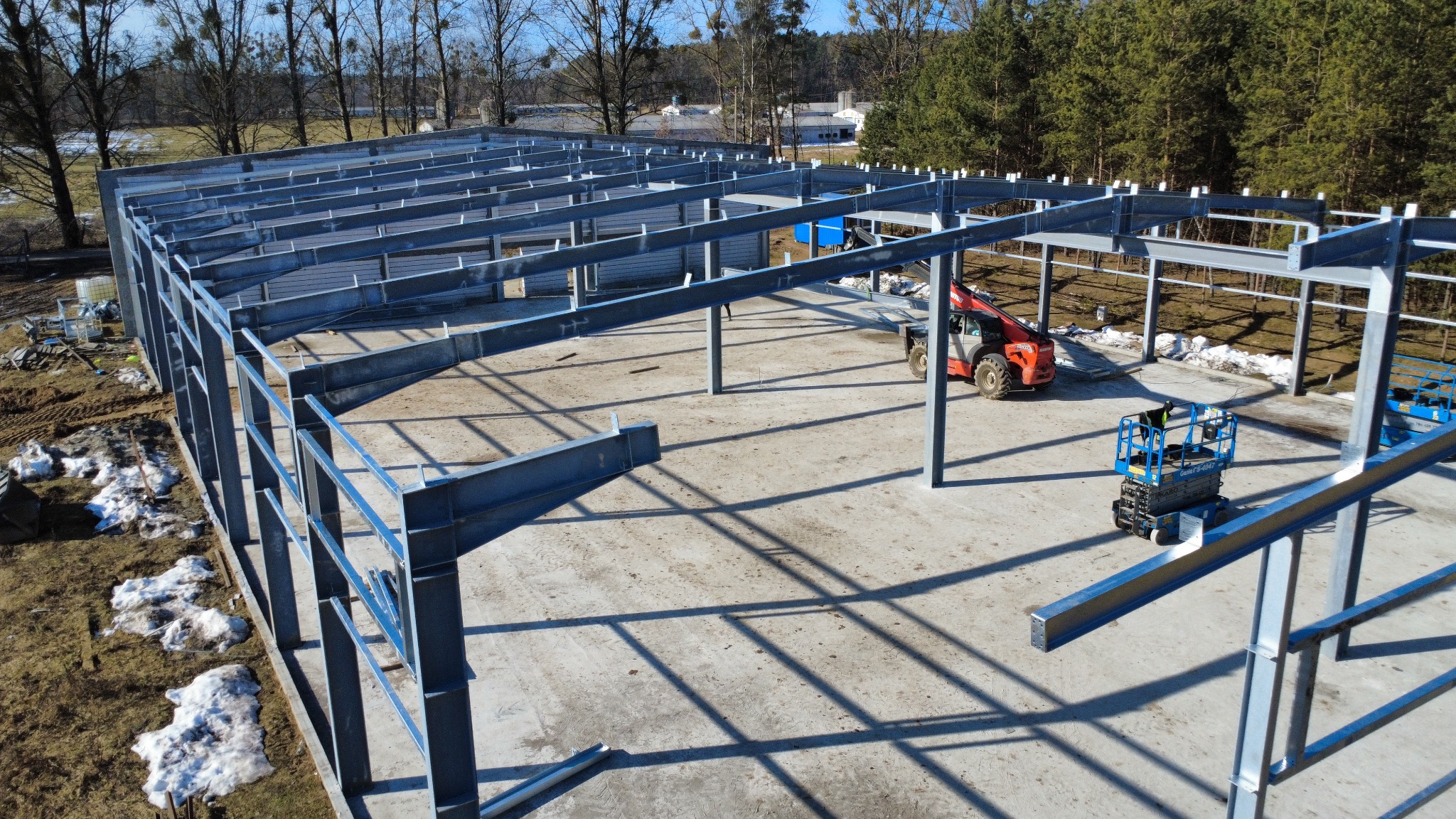 Stalowa konstrukcja hali w trakcie montażu, widok z góry. Na betonowej płycie stoi podnośnik koszowy i czerwona ładowarka teleskopowa. W tle las i budynki.