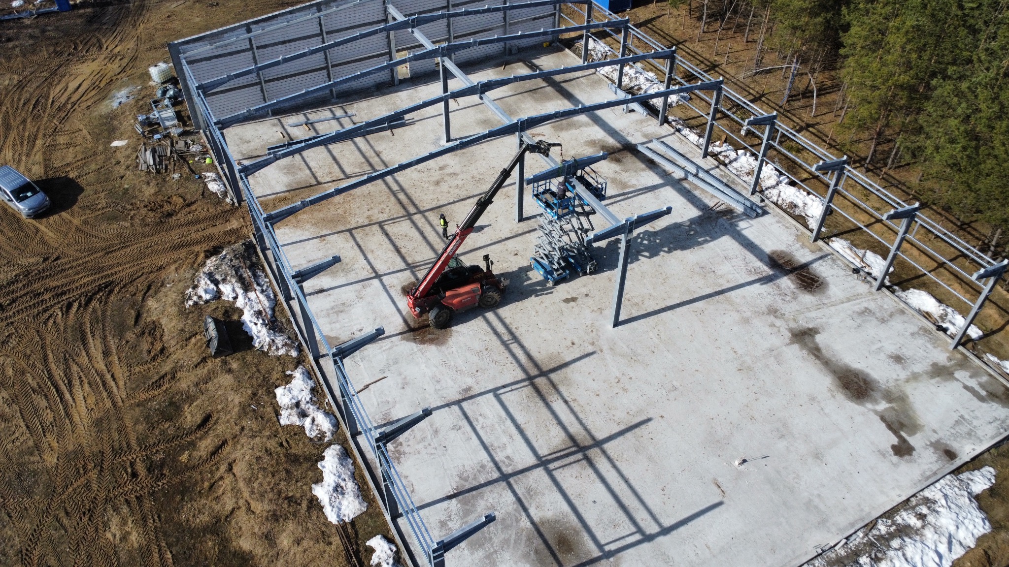 Montaż stalowej konstrukcji hali z użyciem podnośnika teleskopowego na betonowej płycie fundamentowej, widok z góry, częściowo ośnieżony teren.