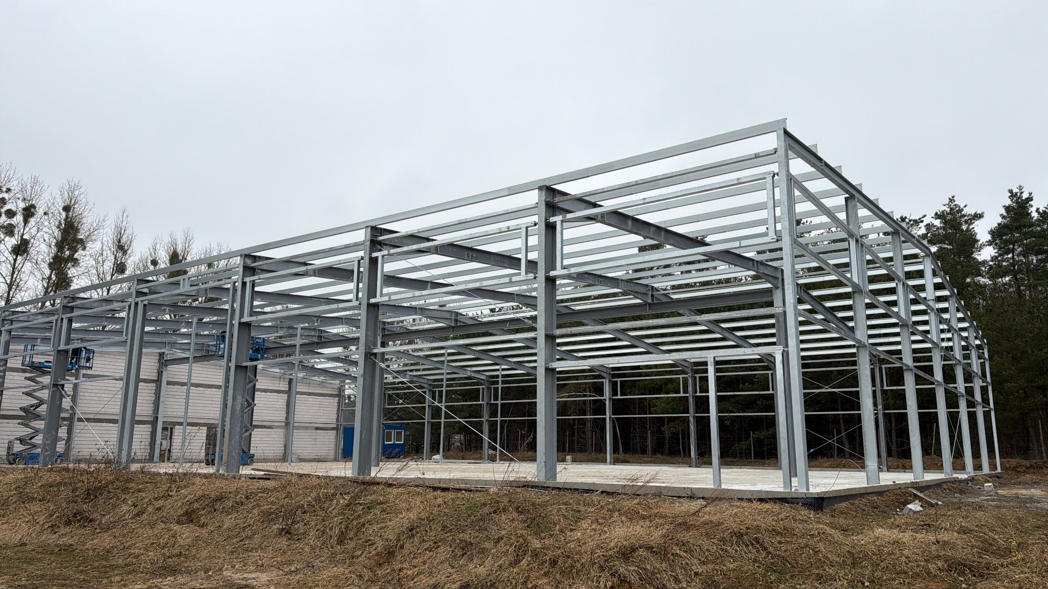 Stalowa konstrukcja hali w trakcie budowy, widoczne podnośniki koszowe, fundamenty i częściowo wymurowane ściany. Tło stanowią drzewa i pochmurne niebo.