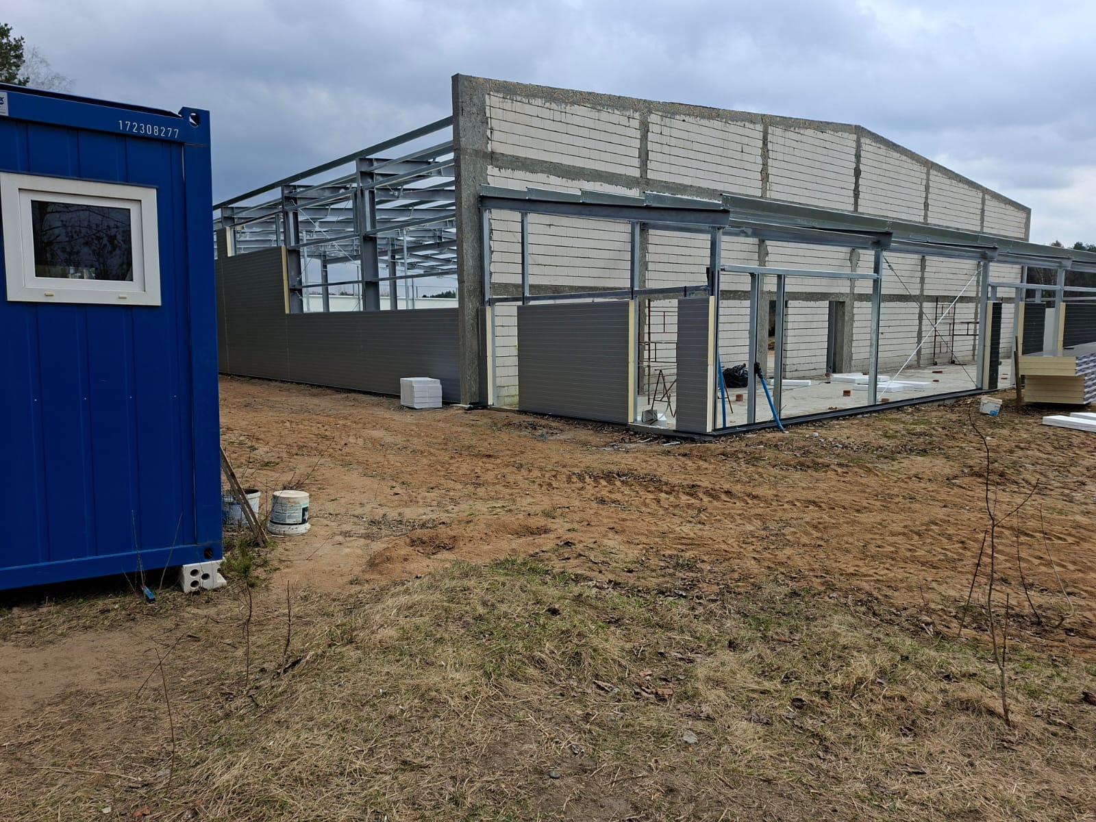 Budowa hali: stalowa konstrukcja, ściany z bloczków i szare panele. Niebieski kontener budowlany z oknem z lewej strony. Teren budowy, pochmurne niebo.