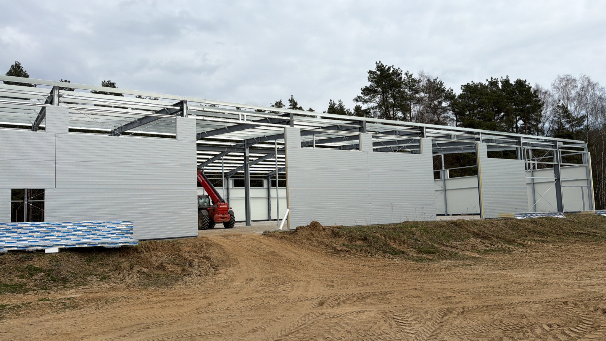 Konstrukcja hali w trakcie montażu: stalowy szkielet, obudowa z paneli warstwowych, widoczny podnośnik teleskopowy i materiały budowlane. Surowy krajobraz, pochmurne niebo.