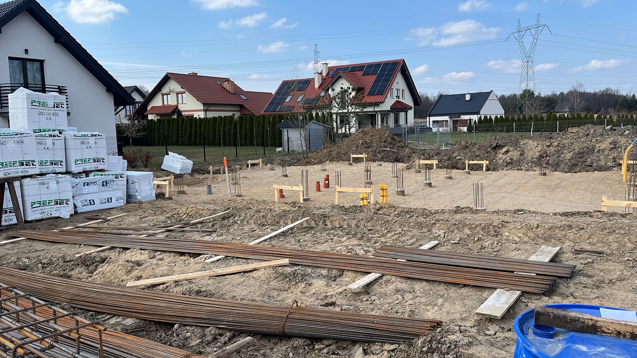 Fundamenty pod dom jednorodzinny: zbrojenie, szalunki, bloczki izolacyjne. Widok na działkę z przygotowanym terenem i sąsiednimi domami w tle.