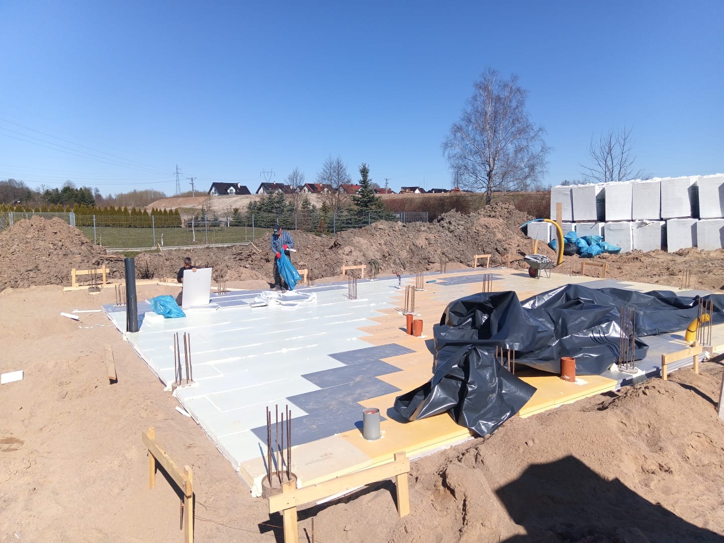 Fundamenty domu jednorodzinnego w trakcie izolacji termicznej, widoczne zbrojenia i przygotowane bloczki. Prace ziemne w toku, słoneczny dzień.