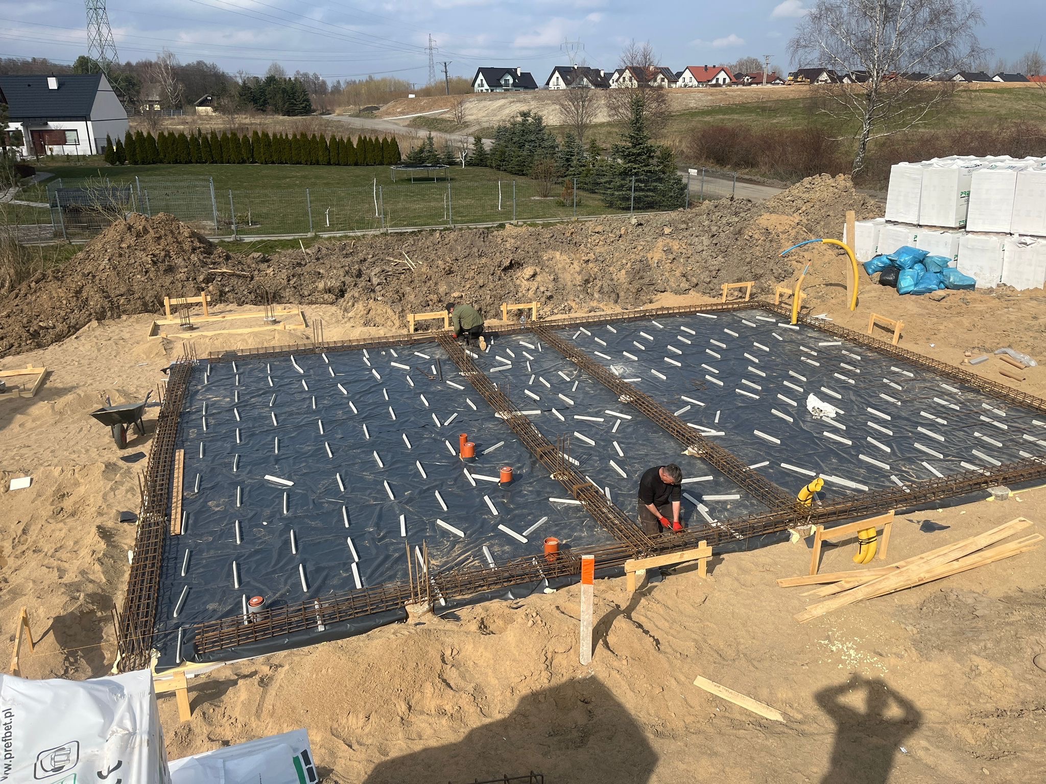 Zbrojenie fundamentów pod budowę domu jednorodzinnego. Widoczne stalowe pręty, folia izolacyjna i białe podkładki dystansowe. W tle zabudowa domów i drzewa.