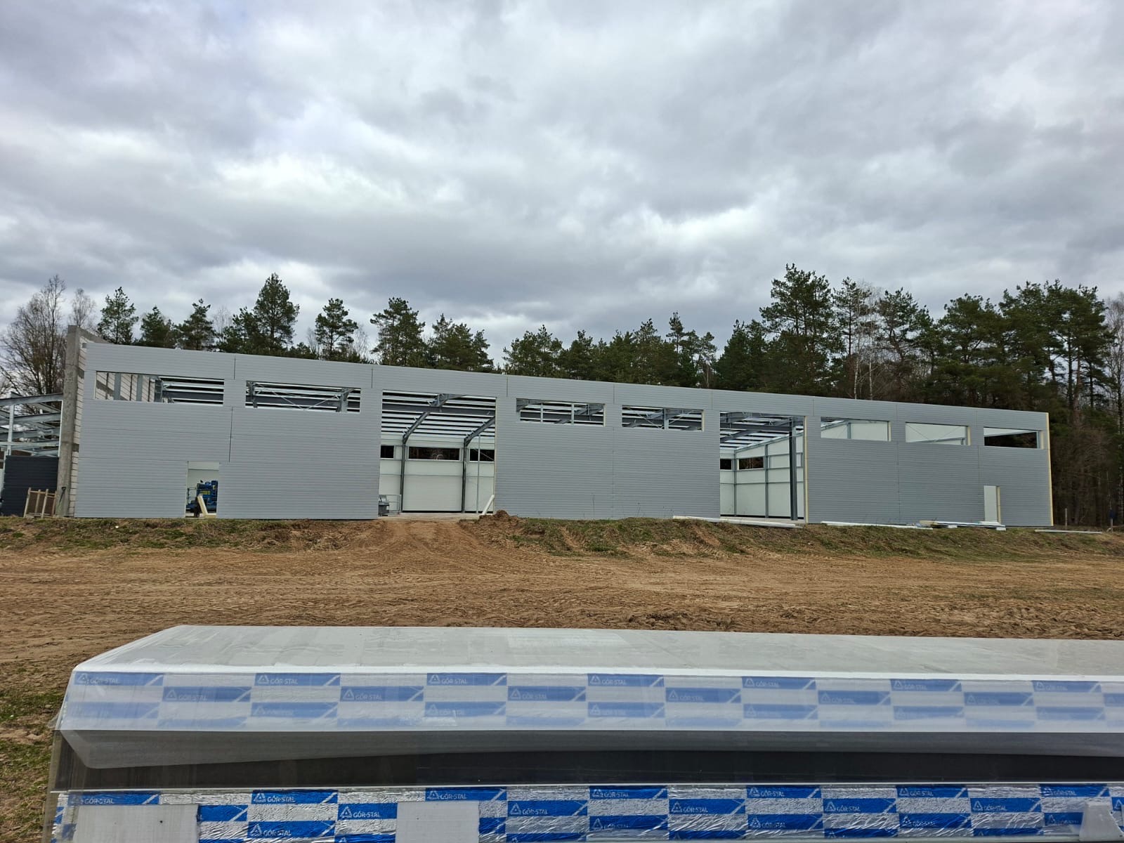 Budowa hali z szarej blachy trapezowej w stanie surowym otwartym, widok z zewnątrz na tle lasu i pochmurnego nieba. Na pierwszym planie elementy konstrukcyjne.