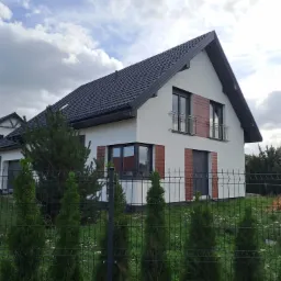 Nowoczesny dom jednorodzinny z białą elewacją, czarną dachówką i drewnianymi akcentami na fasadzie, widziany zza ogrodzenia z tujami.