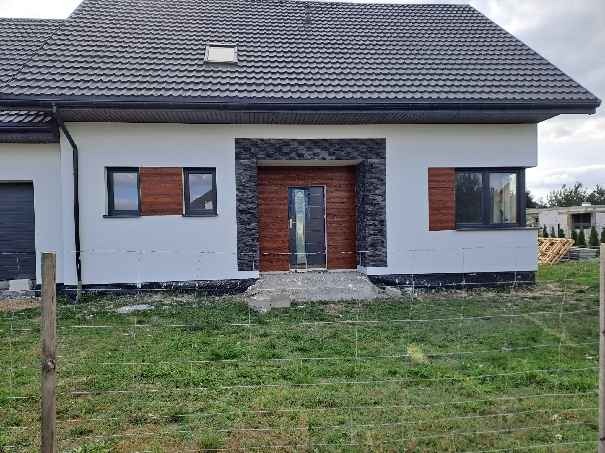 Nowoczesny dom jednorodzinny z białą elewacją, grafitową dachówką i drewnianymi akcentami przy oknach, wykończenie wejścia z szarego kamienia dekoracyjnego, widoczne prace wykończeniowe wokół budynku.