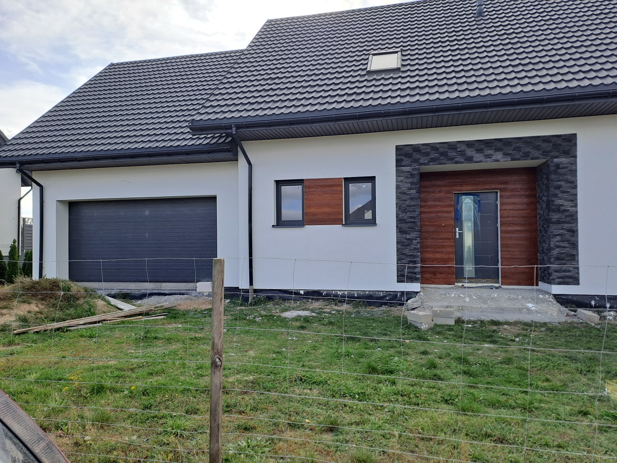 Nowoczesny dom jednorodzinny z białą elewacją, grafitową blachodachówką i garażem, drewnianymi akcentami przy oknach i wejściu oraz elementami dekoracyjnymi z kamienia elewacyjnego. Teren wokół...