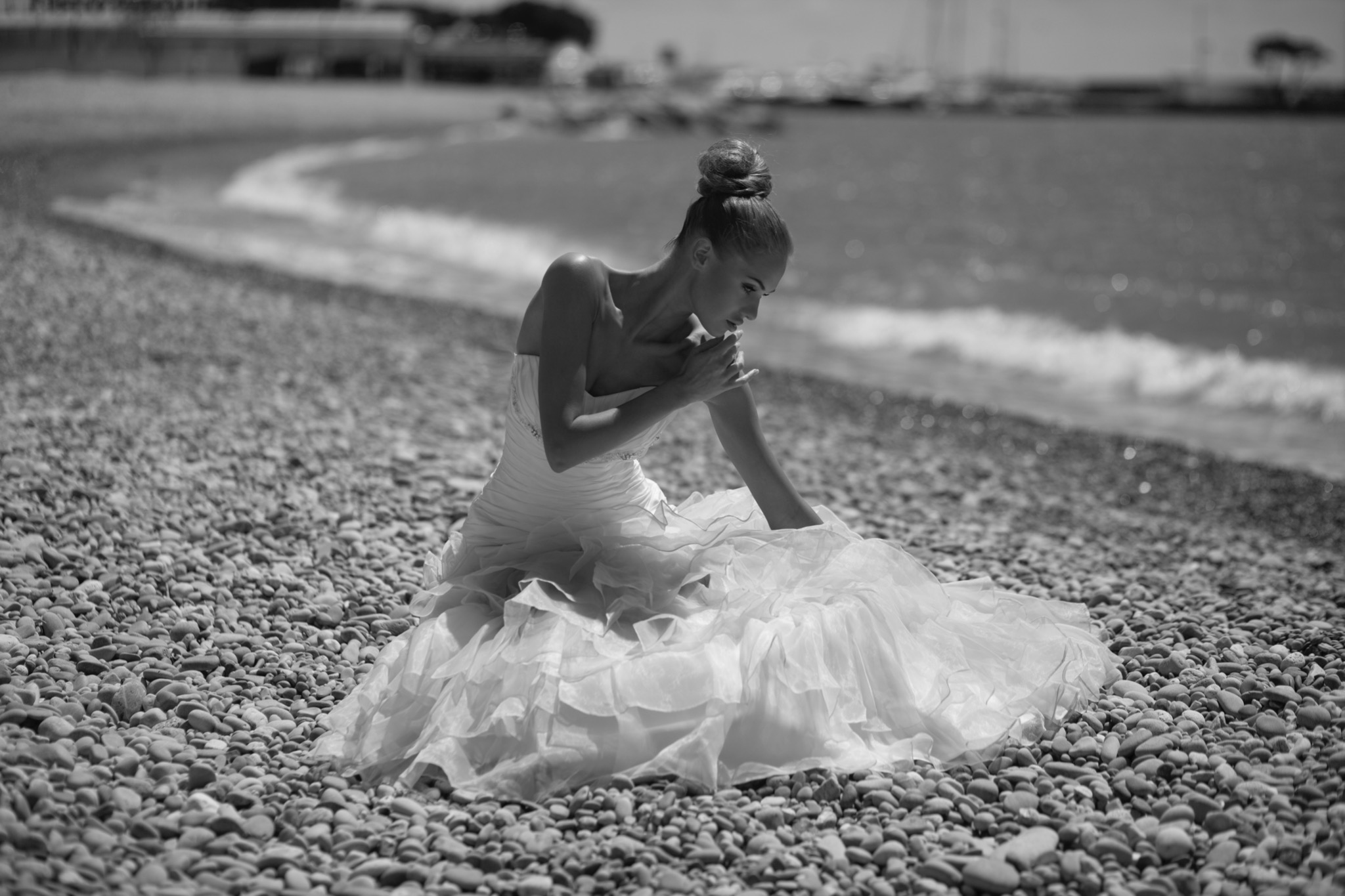 Czarno-białe zdjęcie kobiety w sukni balowej siedzącej na kamienistej plaży z morzem w tle, pochylona głowa, włosy upięte w kok.