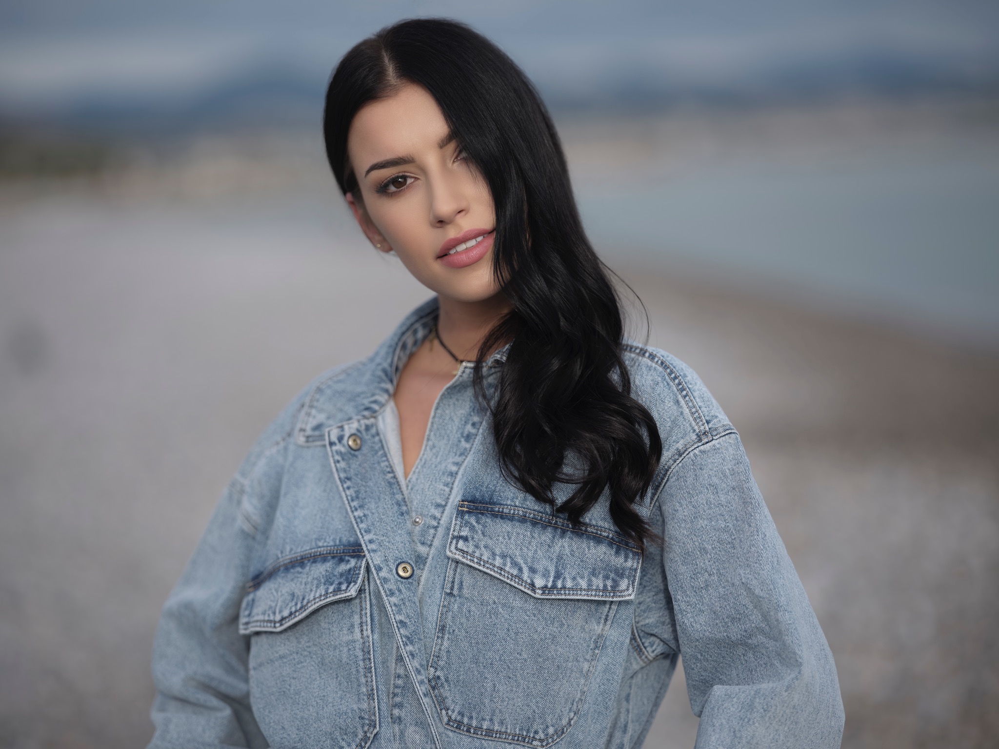 Portret młodej kobiety z długimi, czarnymi włosami w dżinsowej kurtce na tle rozmytego krajobrazu plażowego.
