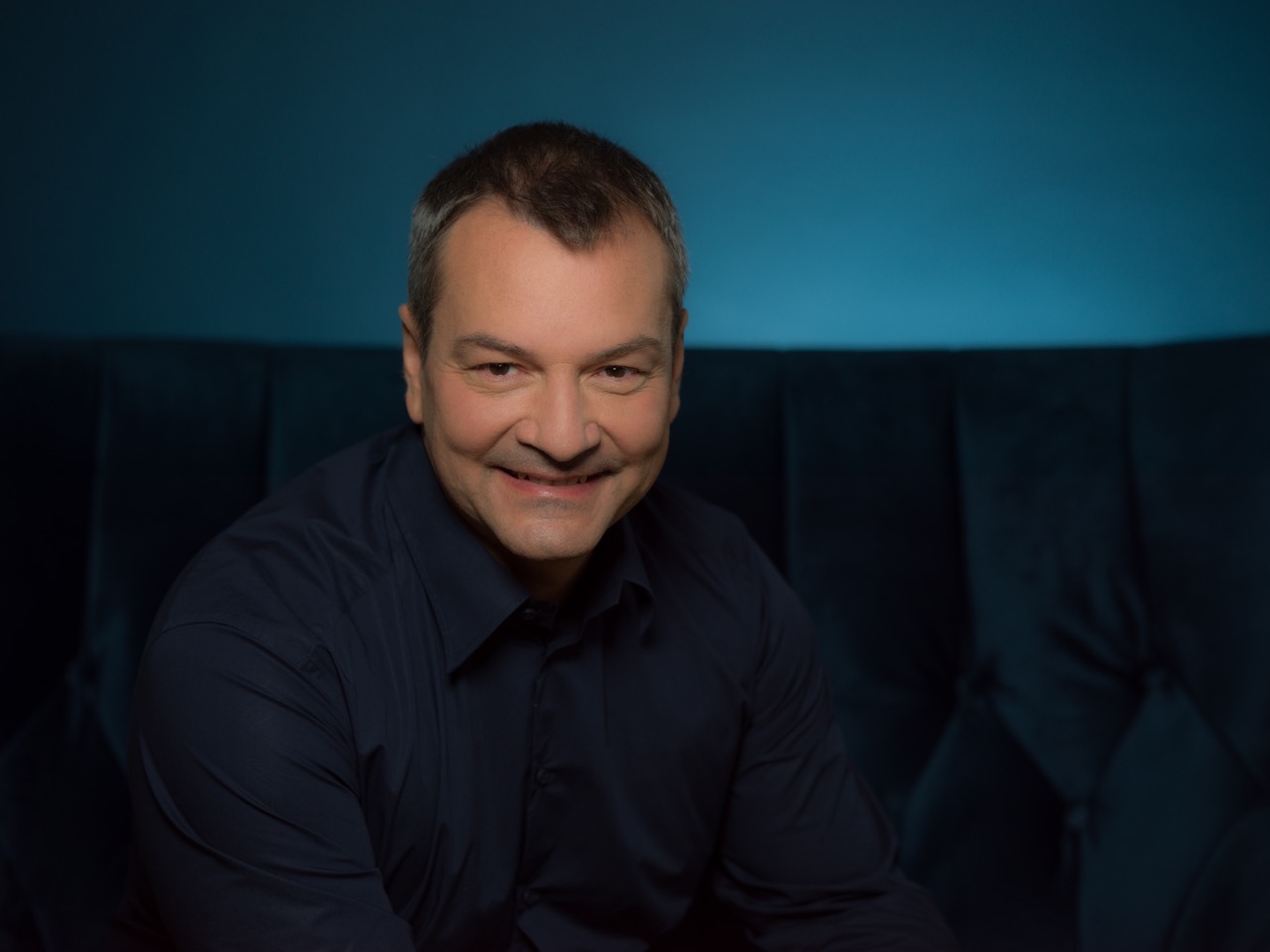 Portret uśmiechniętego mężczyzny w ciemnej koszuli na tle turkusowej ściany i ciemnozielonej kanapy.