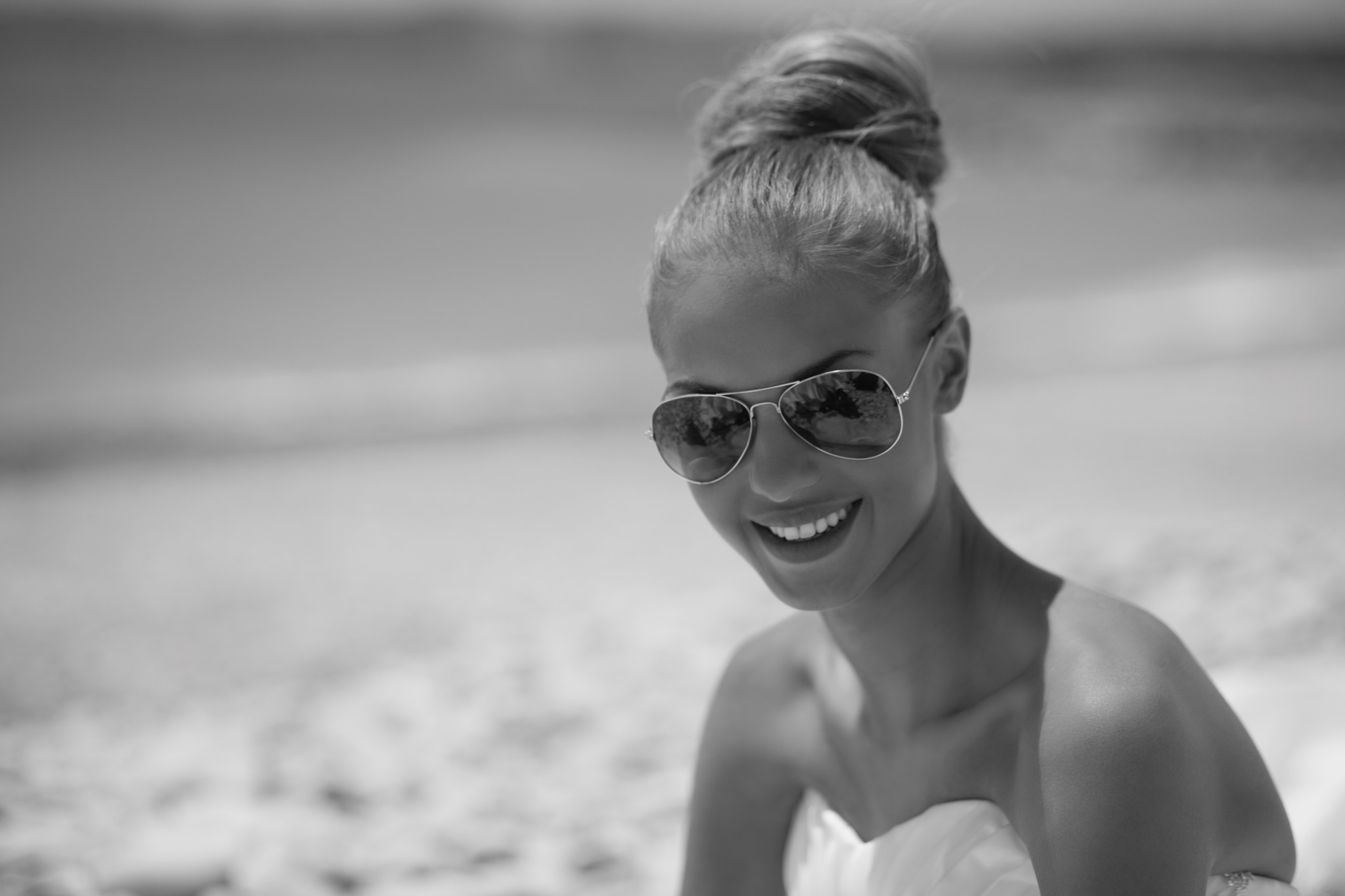 Portret uśmiechniętej kobiety w okularach przeciwsłonecznych na plaży, z włosami upiętymi w kok, w sukni bez ramiączek, czarno-białe zdjęcie.