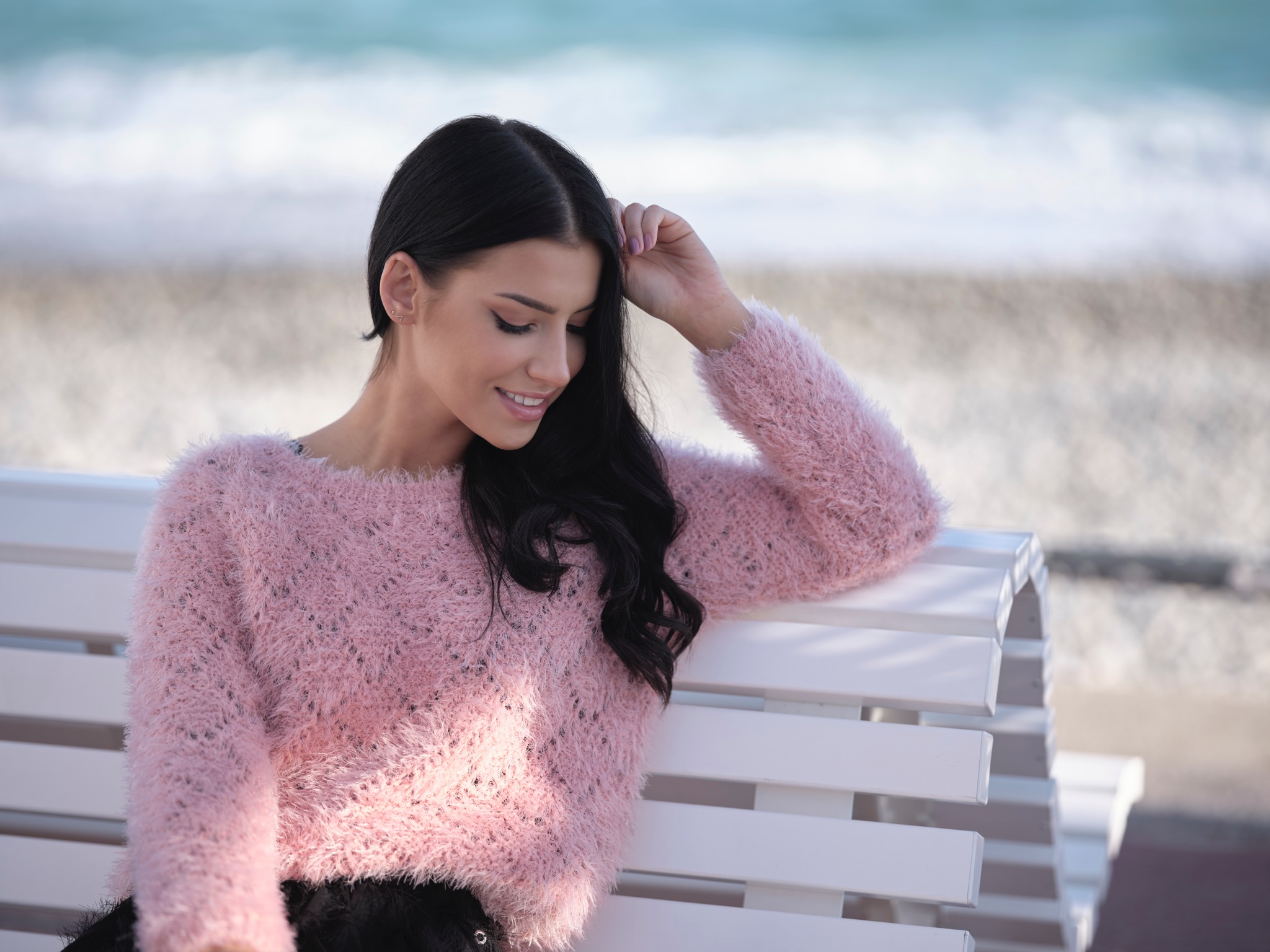 Portret uśmiechniętej, młodej kobiety z ciemnymi włosami w różowym swetrze, siedzącej na białej ławce z widokiem na morze.