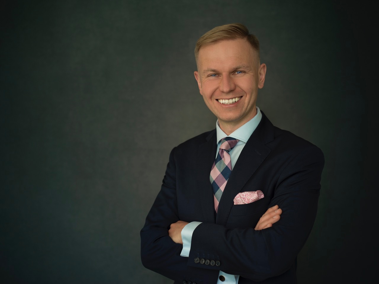 Portret uśmiechniętego mężczyzny w ciemnym garniturze, jasnoniebieskiej koszuli i krawacie w kratę z różowymi akcentami, z chusteczką w kieszonce, na ciemnoszarym tle.