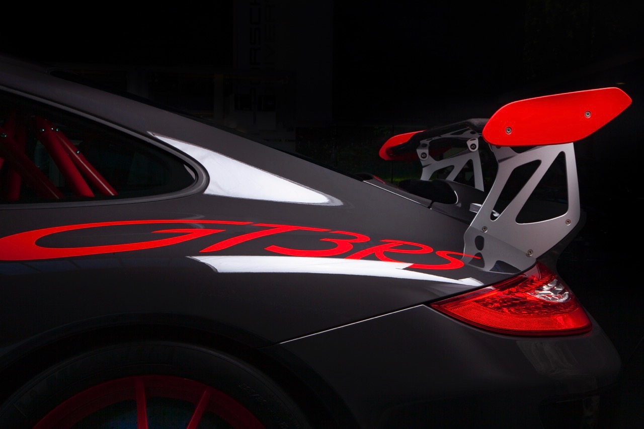 Szary samochód sportowy Porsche GT3 RS z czerwonymi akcentami, widoczny tylny spojler, felgi i napisy, sfotografowany w studiu z ciemnym tłem.