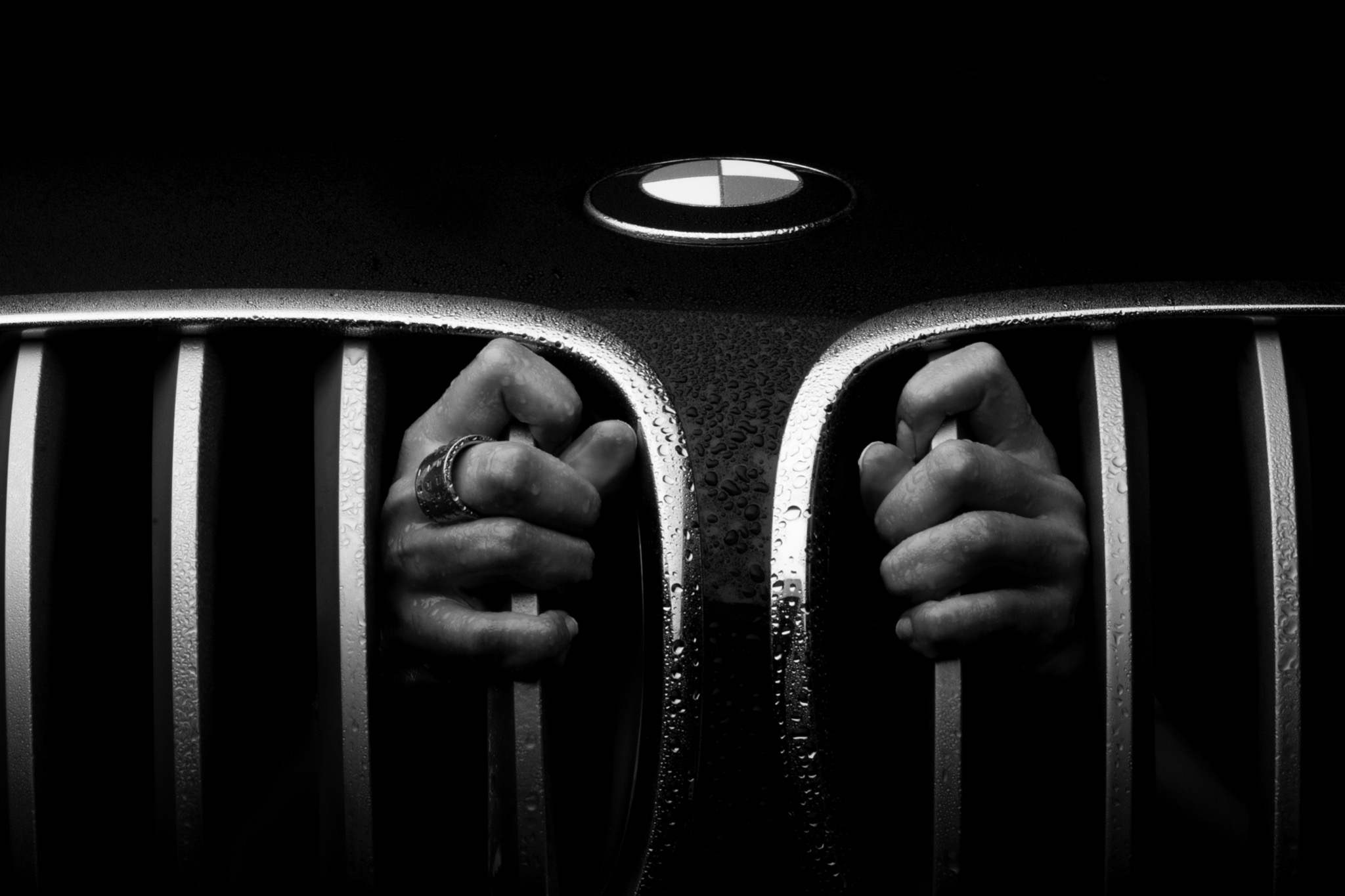 Czarno-białe zdjęcie detalu przedniej atrapy samochodu BMW z mokrymi prętami, za którymi widać dłonie mężczyzny w pierścieniu.