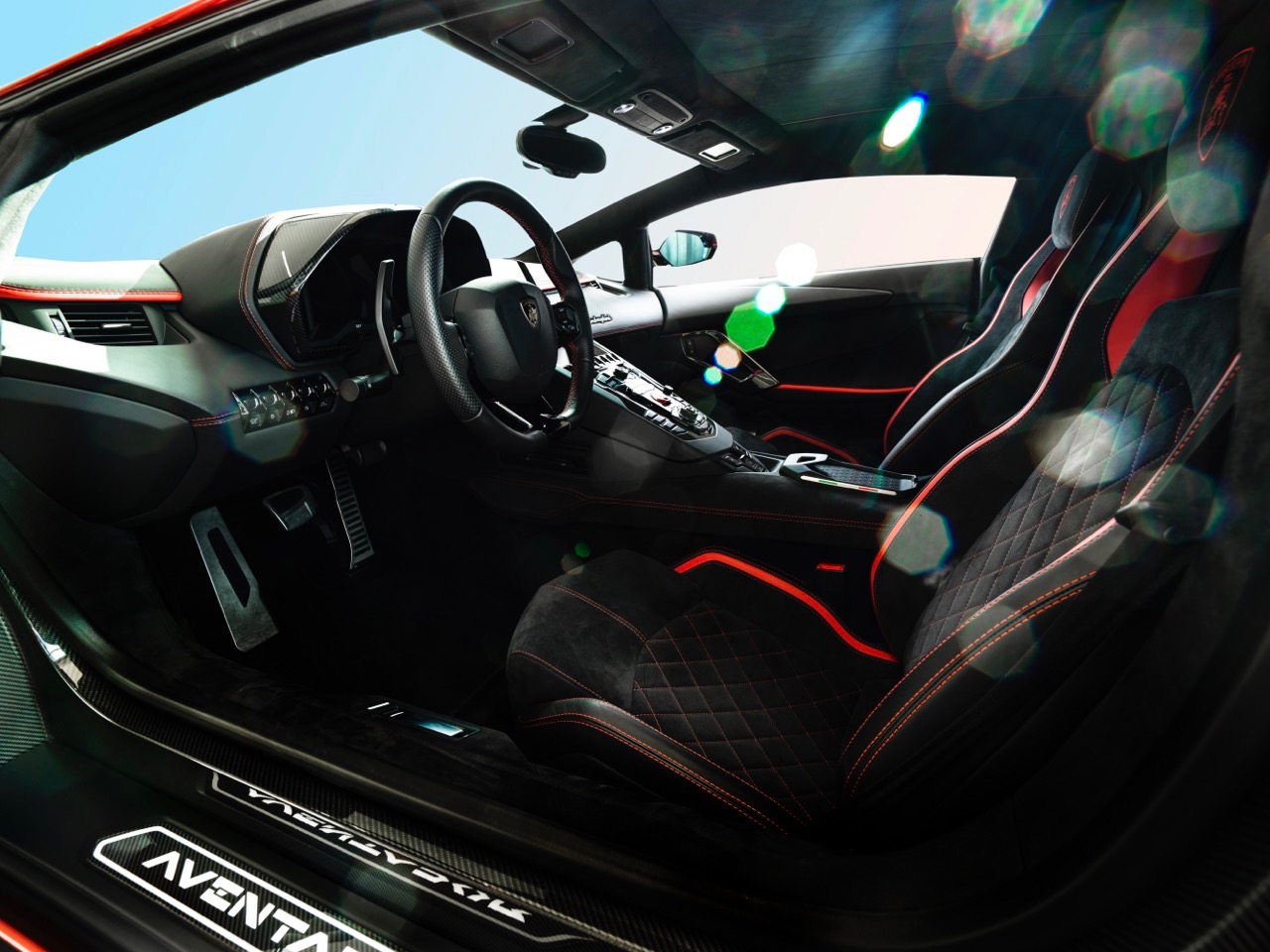 Wnętrze sportowego samochodu Lamborghini Aventador z czarną tapicerką, czerwonymi akcentami i karbonowymi detalami, widoczne fotele z pikowaniem i logo na zagłówkach.