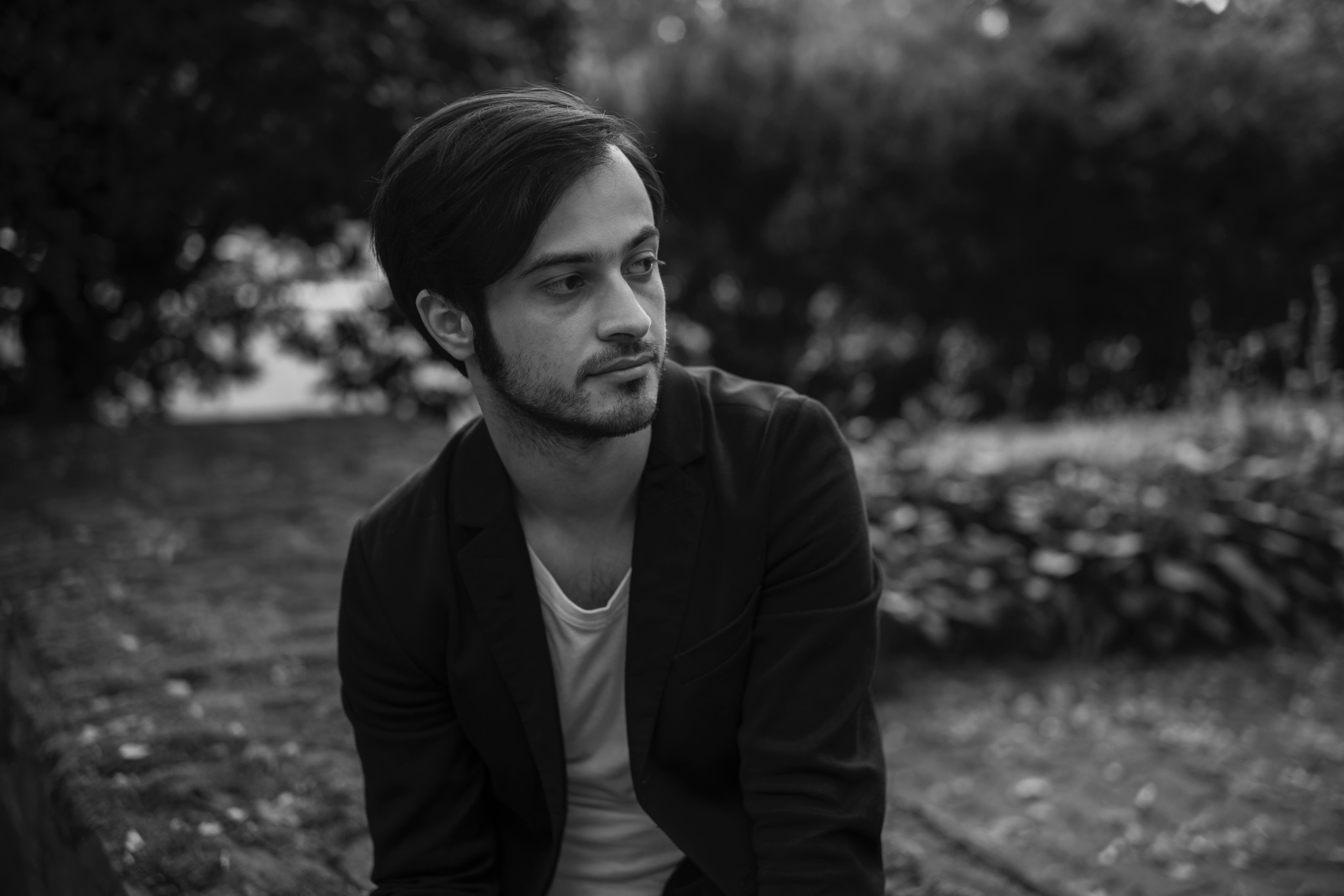 Portret młodego mężczyzny w marynarce, z delikatnym zarostem, spoglądającego w bok na tle rozmytej zieleni w czerni i bieli.