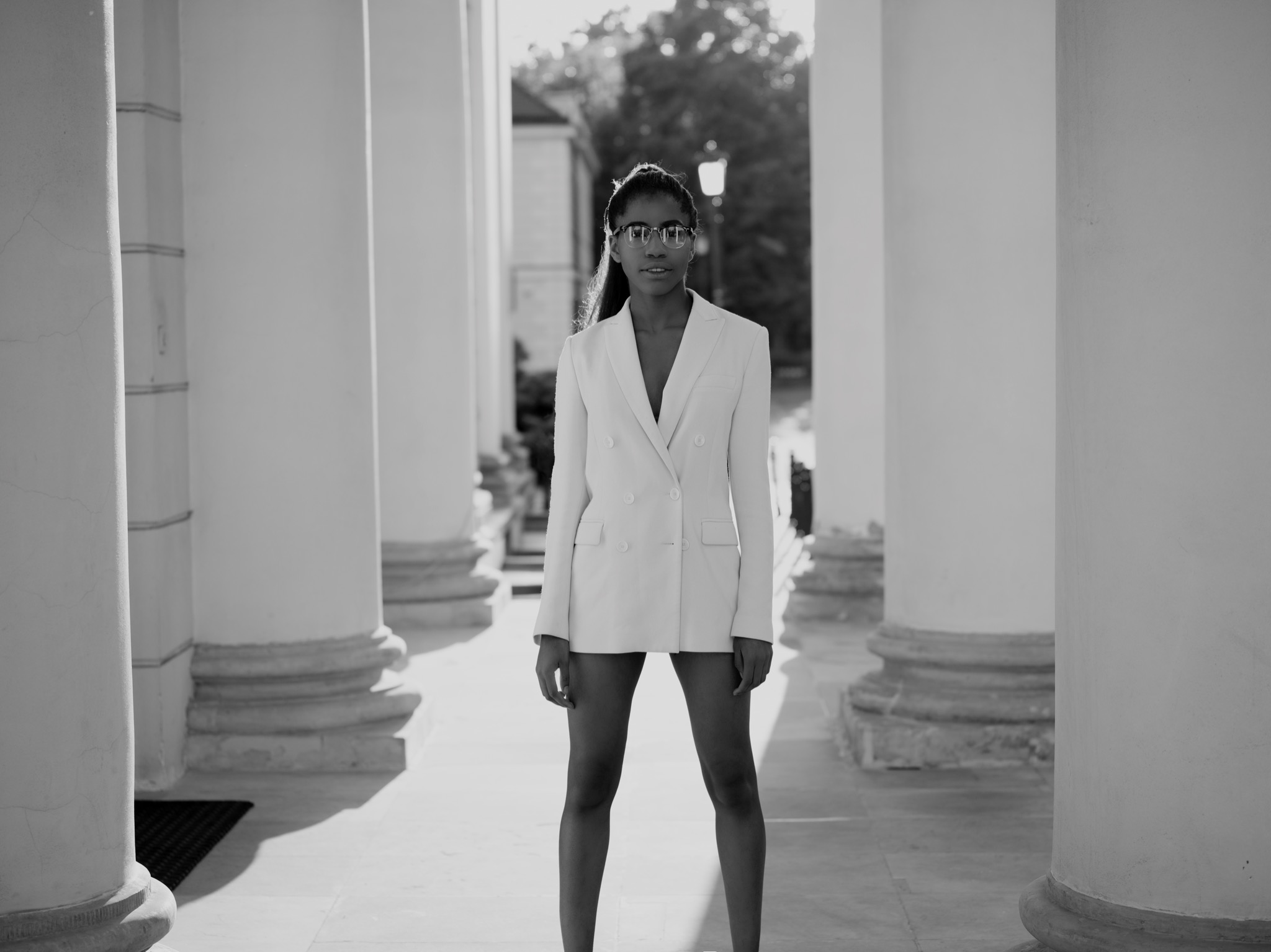 Czarno-białe zdjęcie kobiety w okularach i białym garniturze, stojącej między kolumnami z widocznymi promieniami słonecznymi.