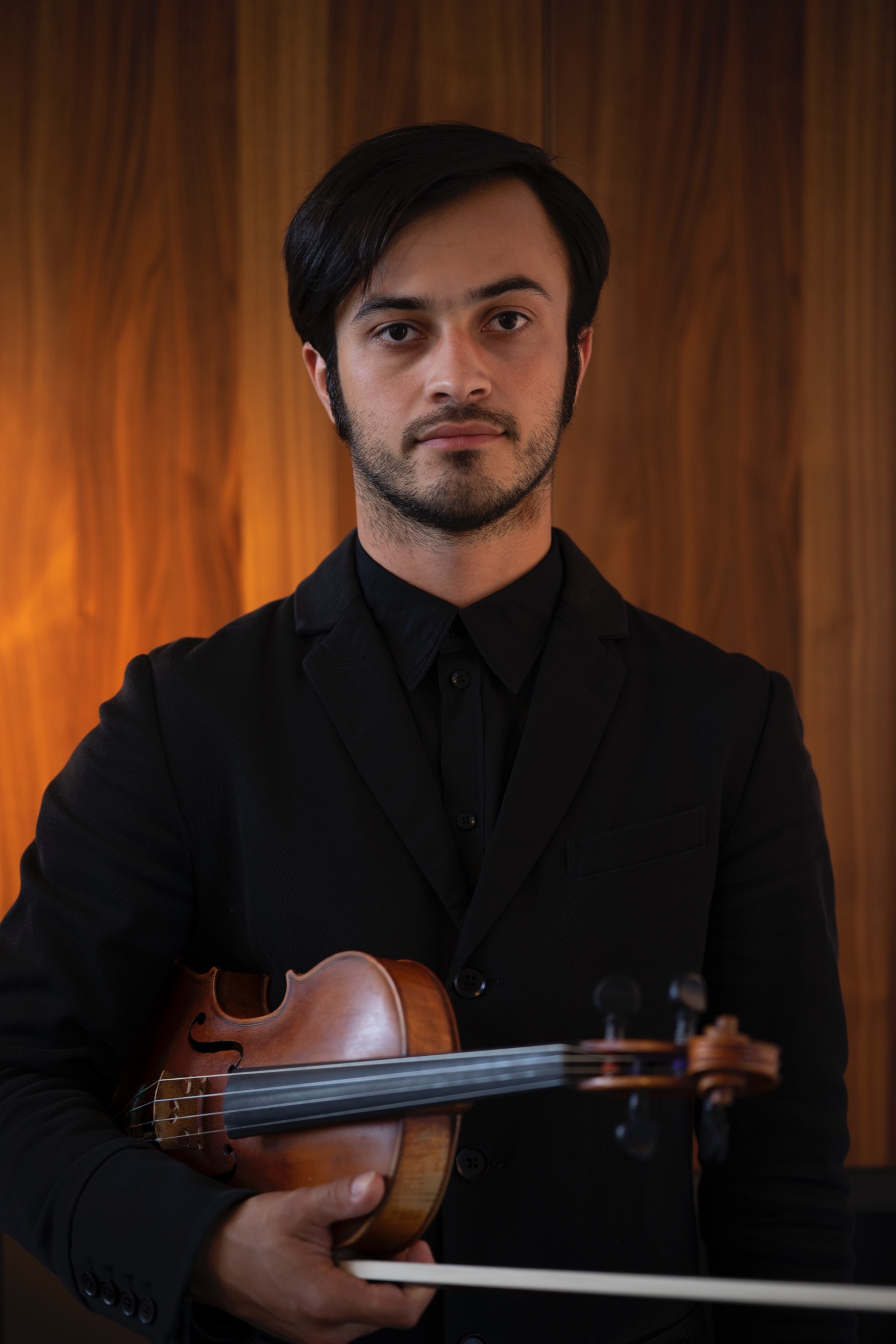 Portret młodego mężczyzny w czarnym garniturze, trzymającego skrzypce na tle drewnianej ściany.