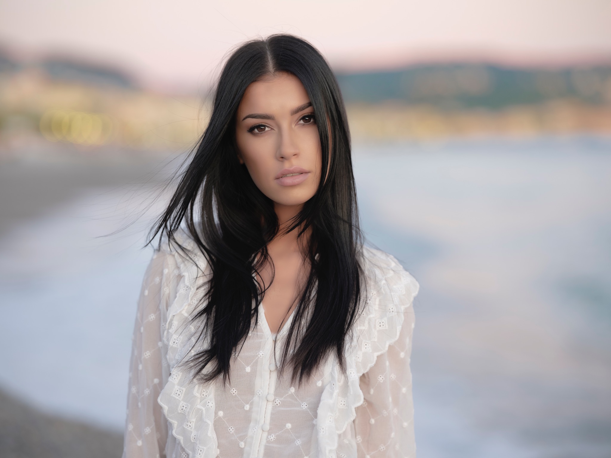 Portret młodej kobiety o długich, czarnych włosach w białej, koronkowej bluzce na tle rozmytej plaży o zachodzie słońca.
