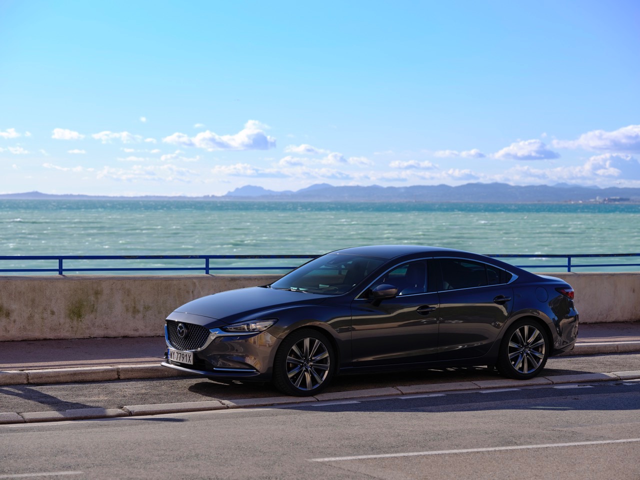 Ciemnoszary samochód marki Mazda zaparkowany przy nadmorskiej promenadzie z widokiem na błękitne morze i górzysty horyzont pod jasnym, błękitnym niebem.