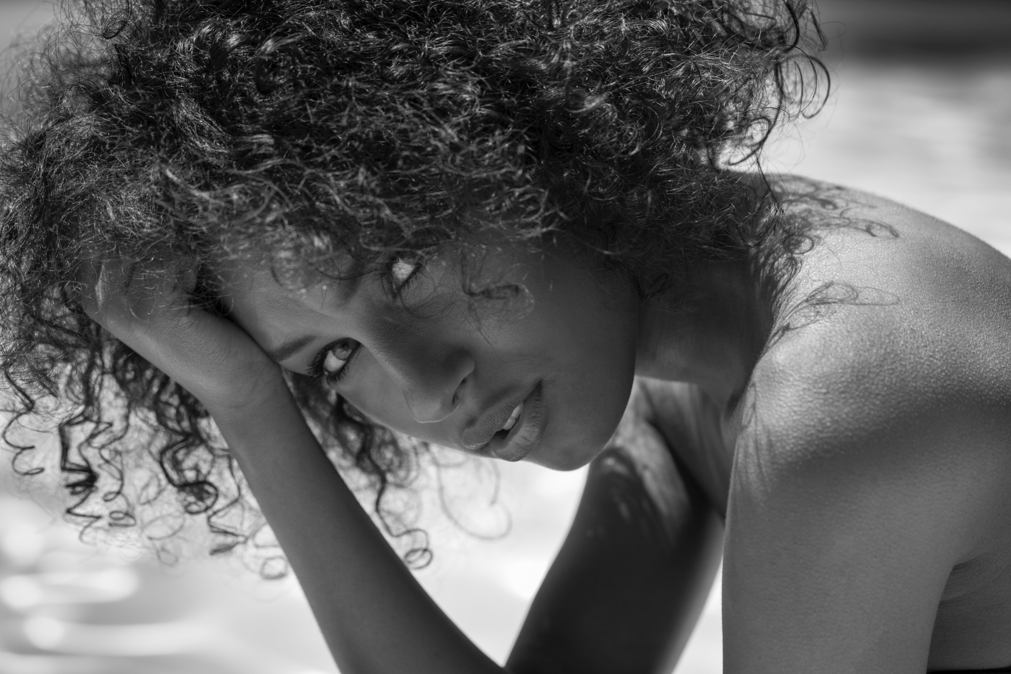 Portret kobiety z kręconymi włosami, opierającej głowę na dłoni, w czarno-białej tonacji, z widocznym tatuażem na ramieniu i delikatnym makijażem.
