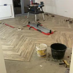 Układanie parkietu w jodełkę z płytek imitujących drewno, widoczny sprzęt glazurniczy, wiaderka, worek z zaprawą i poziomica.