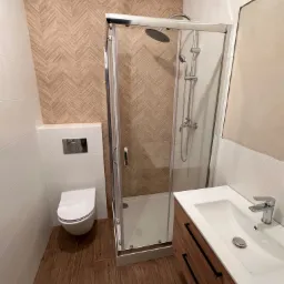 Nowoczesna łazienka z kabiną prysznicową, umywalką z szafką z drewnianymi frontami i toaletą podwieszaną. Ściany wyłożone białymi i drewnopodobnymi płytkami w jodełkę, podłoga z płytek imitujących...